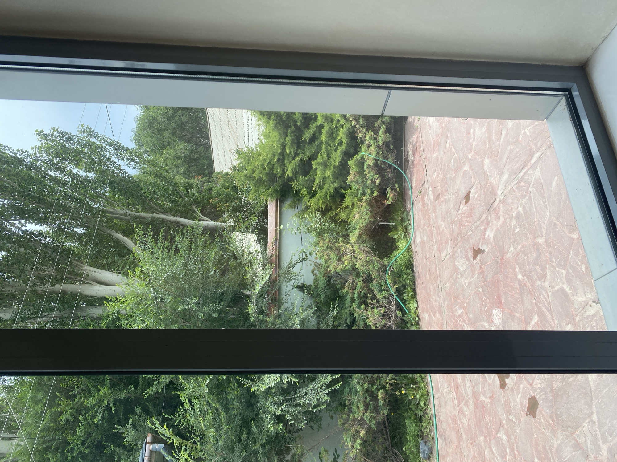This image, taken from inside a building in Balykchy, Kyrgyzstan, shows a view of a backyard through a large window. The backyard features a paved patio made of reddish-brown irregular stones. Beyond the patio, there is lush greenery with various types of bushes and trees. A green garden hose is visible coiled on the ground amongst the foliage. A light-colored, textured wall runs along the back of the yard, partially obscured by the dense vegetation. The sky above is partly cloudy. The framing of the image includes a dark gray window frame on the left and a lighter beige wall on the right, indicating the perspective is from an indoor setting looking outwards.