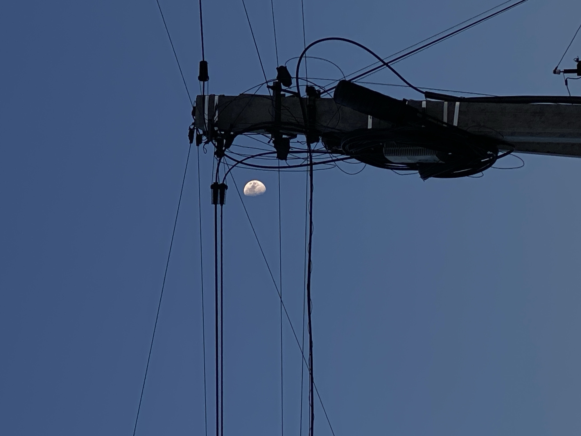 A concrete utility pole, adorned with numerous black wires and cables, stands prominently in the foreground. The pole is a dark gray, rough-textured column, its surface crisscrossed by a dense network of electrical and telecommunication lines. The wires stretch across the frame, some taut and straight, others forming gentle arcs. Attached to the pole are various pieces of equipment, including what appear to be junction boxes and insulators, also shrouded in bundles of dark cabling.
To the left of the utility pole, a crescent moon is visible in the clear, deep blue sky. The moon is illuminated on its right side, with visible craters and surface details. The sky itself is a uniform, cloudless blue, suggesting a twilight or early evening setting. The overall impression is one of a utilitarian urban landscape juxtaposed with a celestial body. The scene is likely located in Cairaclia, Moldova, given the context of the request. No people or animals are present in the image.