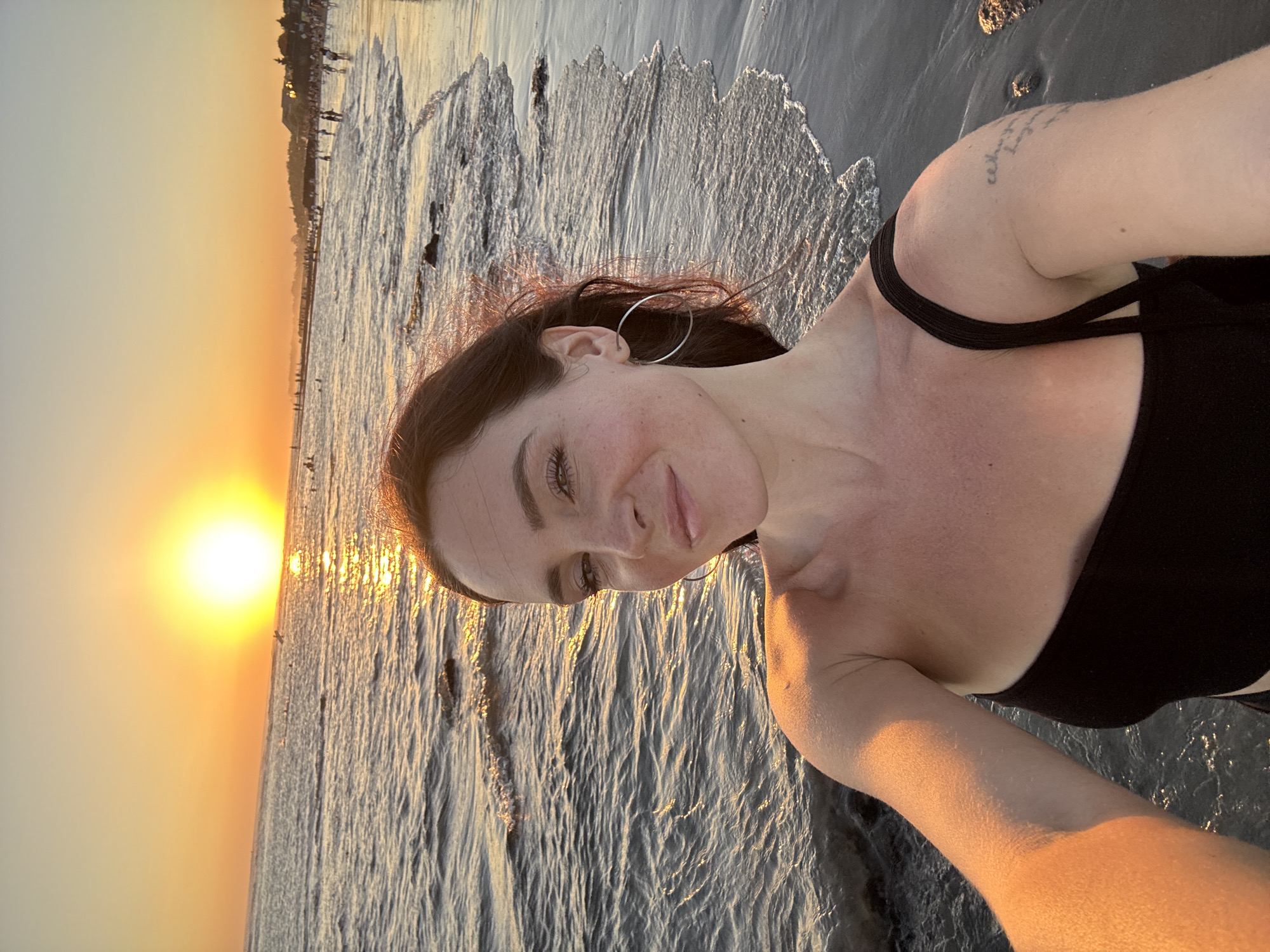 A woman is taking a selfie on a beach at sunset. The sun is setting over the ocean, casting a warm glow on the scene. The woman is wearing a black swimsuit and has her hair down. She is smiling and looking at the camera. There are people in the background, some of whom are walking along the shore and others are in the water. The beach is dark sand, and the water is calm. The location is North Kuta, Indonesia.