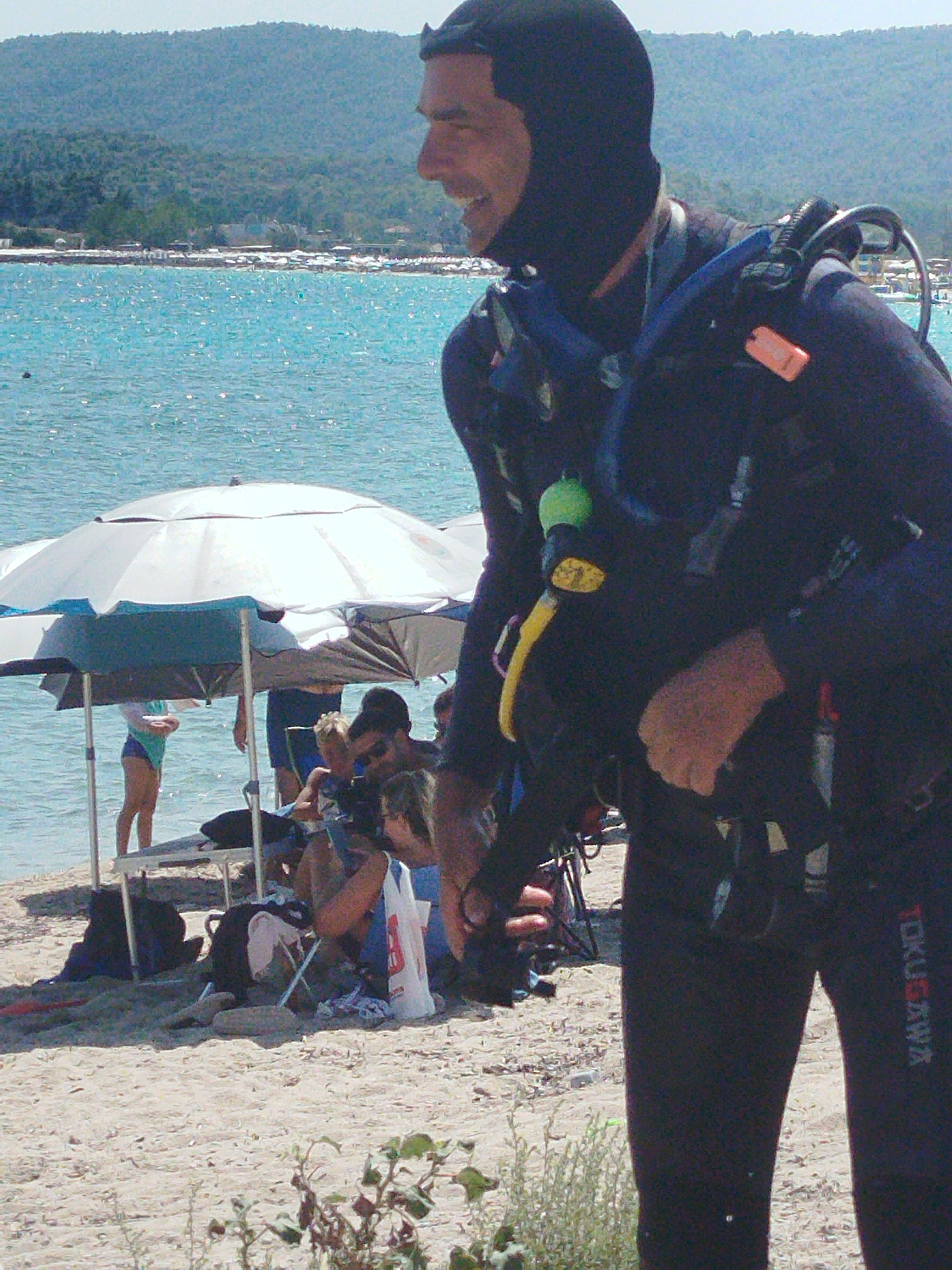 A man in a black full-body wetsuit and hood stands on a sandy beach in Sithonia, Greece, facing right. He wears a scuba diving mask and regulator. The man's right arm is raised and bent, and he is looking to his right, smiling. Behind him, the turquoise sea stretches to the horizon, with a hilly, tree-covered shoreline in the distance. Several beach umbrellas and a few people are visible in the background, some sitting and some standing. One person is wearing a light blue swimsuit. The scene appears to be a sunny day at the beach.