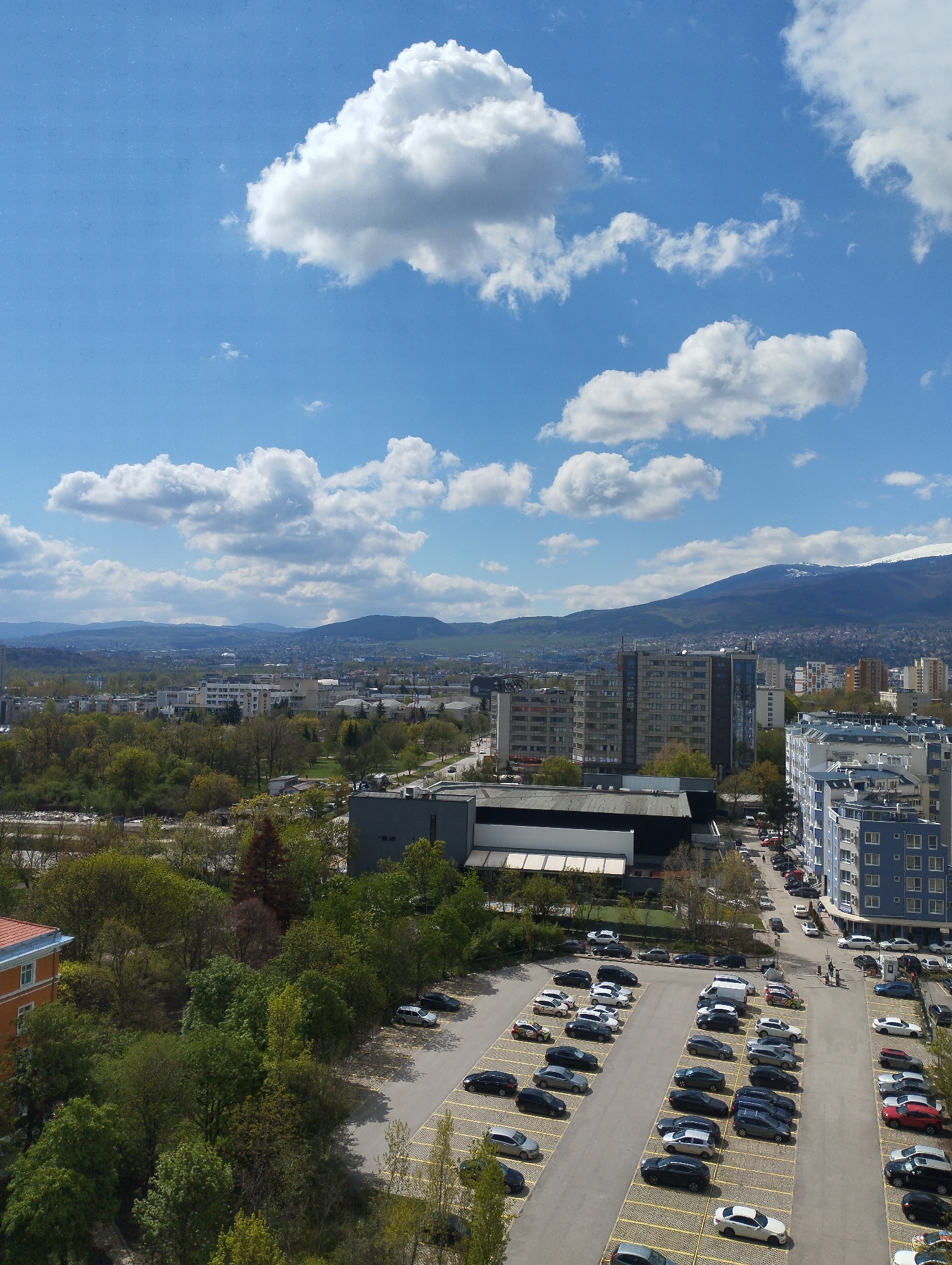 This high-angle, panoramic image captures a vibrant urban landscape under a bright, partly cloudy sky. Fluffy white cumulus clouds are scattered across the vivid blue sky, which transitions to a lighter hue near the horizon. In the distant background, a range of mountains is visible, with one prominent peak partially covered in snow, suggesting a cooler season or higher altitude.
The mid-ground of the image features a sprawling city with a mix of buildings of various heights and architectural styles, interspersed with numerous green spaces and trees. Several multi-story residential and commercial buildings stand out, including a distinctive blue-sided apartment block with white details on the right and taller, more modern structures in the center-right. Dense clusters of deciduous trees, some in early foliage, indicate parks or wooded areas within the city.
The foreground is dominated by large asphalt parking lots, meticulously marked with yellow lines, and filled with a diverse array of parked cars of different makes, models, and colors. A few roads with some moving vehicles are also visible, along with small, indistinct figures of people in the bottom right corner, suggesting pedestrian activity. To the far left, a building with an orange roof and blue trim is partially visible amidst the greenery.
The scene is set on a clear, sunny day, indicated by the bright illumination and distinct shadows. The lush green trees suggest spring or early summer. No specific event is occurring; rather, the image captures a moment of everyday city life. There is no discernible text visible in the photograph.