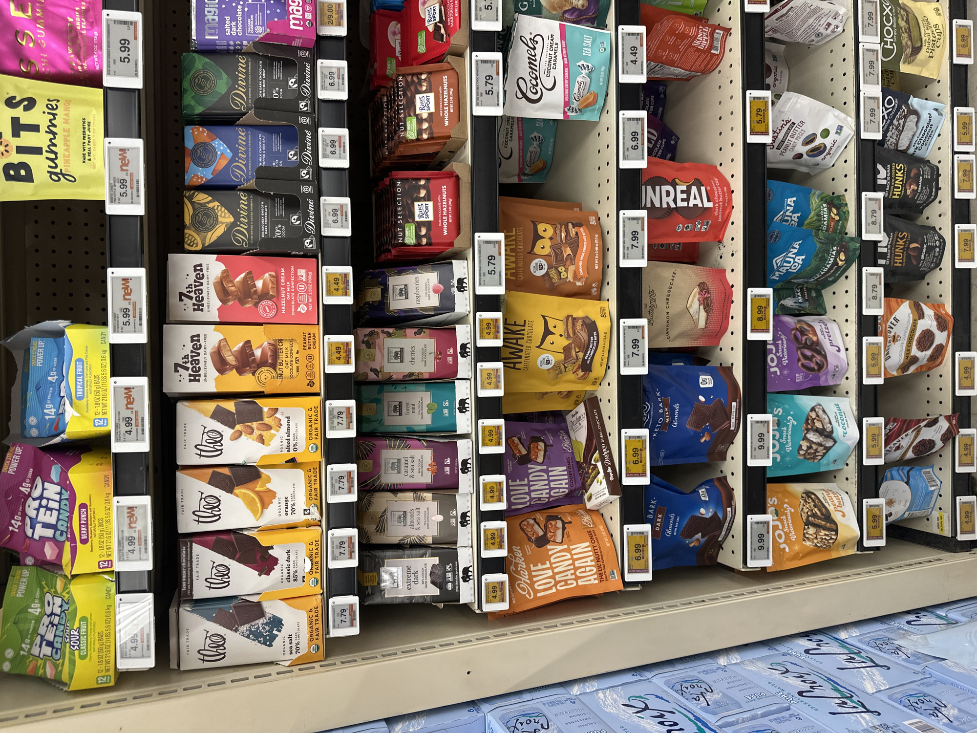 The image shows a shelf display of various chocolate bars and snacks in a retail store. The products are arranged in rows on white shelving units. Prominently featured are brands like Theo, 7th Heaven, Divine, Magic, Harken, Love Candy Again, Awake, Cocomels, JoJo's, Mauna Loa, Justin's, Uncover, Hunks, and HOCXO. Prices are displayed on white tags beneath the products. The scene is well-lit, indicating an indoor retail environment. The location is identified as Schaumburg, United States of America. There are no people visible in the image.