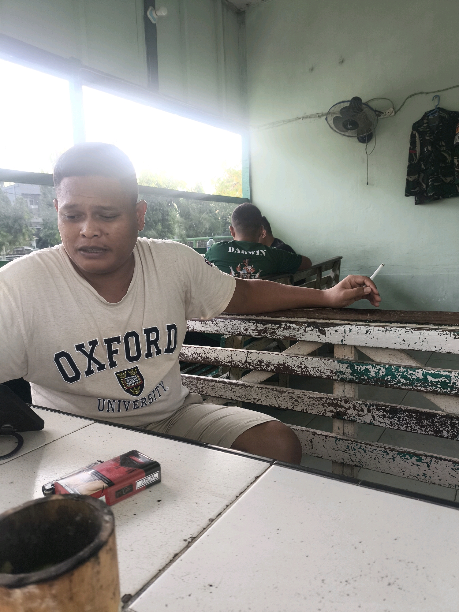 A man wearing a light-colored t-shirt with "OXFORD UNIVERSITY" printed on it is seated at a white table. He is looking to his left and appears to be speaking. To his left on the table is a pack of cigarettes and a cylindrical object. Behind him, another person is visible, wearing a green t-shirt with "DARWIN" printed on it, and holding a lit cigarette. The background shows a wall painted light green, a fan mounted on the wall, and camouflage clothing hanging on a hook. The setting appears to be an outdoor or semi-outdoor covered area, possibly a warung or food stall, characterized by weathered wooden benches and tables. The lighting suggests it is daytime. The image is taken in Gorontalo, Indonesia.
