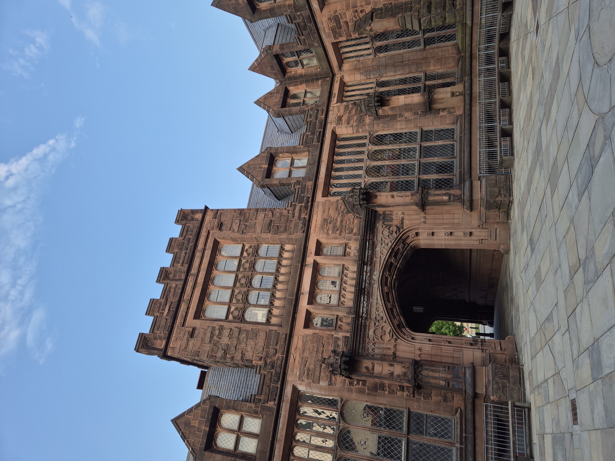 The photograph captures a view of a Gothic Revival building on a clear day in Princeton, New Jersey, United States of America. The building is constructed of reddish-brown stone, featuring crenellated towers, pointed arches, and numerous Gothic-style windows with diamond-shaped panes. A prominent arched entryway is visible in the center of the frame, leading into a dimly lit interior. The exterior is adorned with decorative stonework. In front of the building, a courtyard area is paved with large, irregular flagstones. A metal bench with a stone seat is positioned to the left of the entryway, and a low metal fence with a stone seating area runs along the front of the building to the right. The sky above is a clear blue with a few wispy white clouds. The overall scene suggests an architectural institution, possibly a university campus, given the style and grandeur of the structure. No people are clearly visible or engaged in any discernible activity within the frame.