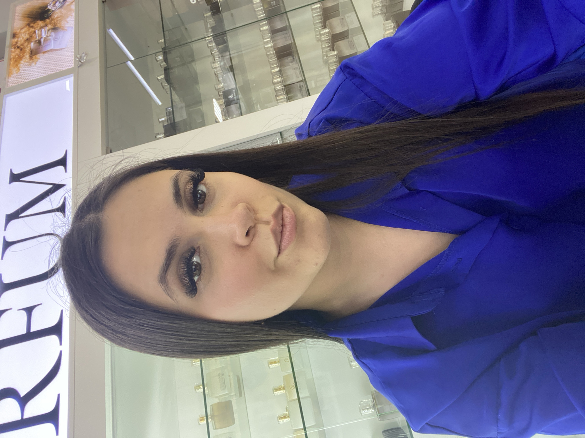 A woman is looking at the camera. She has dark hair and is wearing a blue top. She appears to be standing inside a retail store, with shelves of perfumes and other products visible behind her. The city of Chaikovsky, Russia is not discernible from the image.