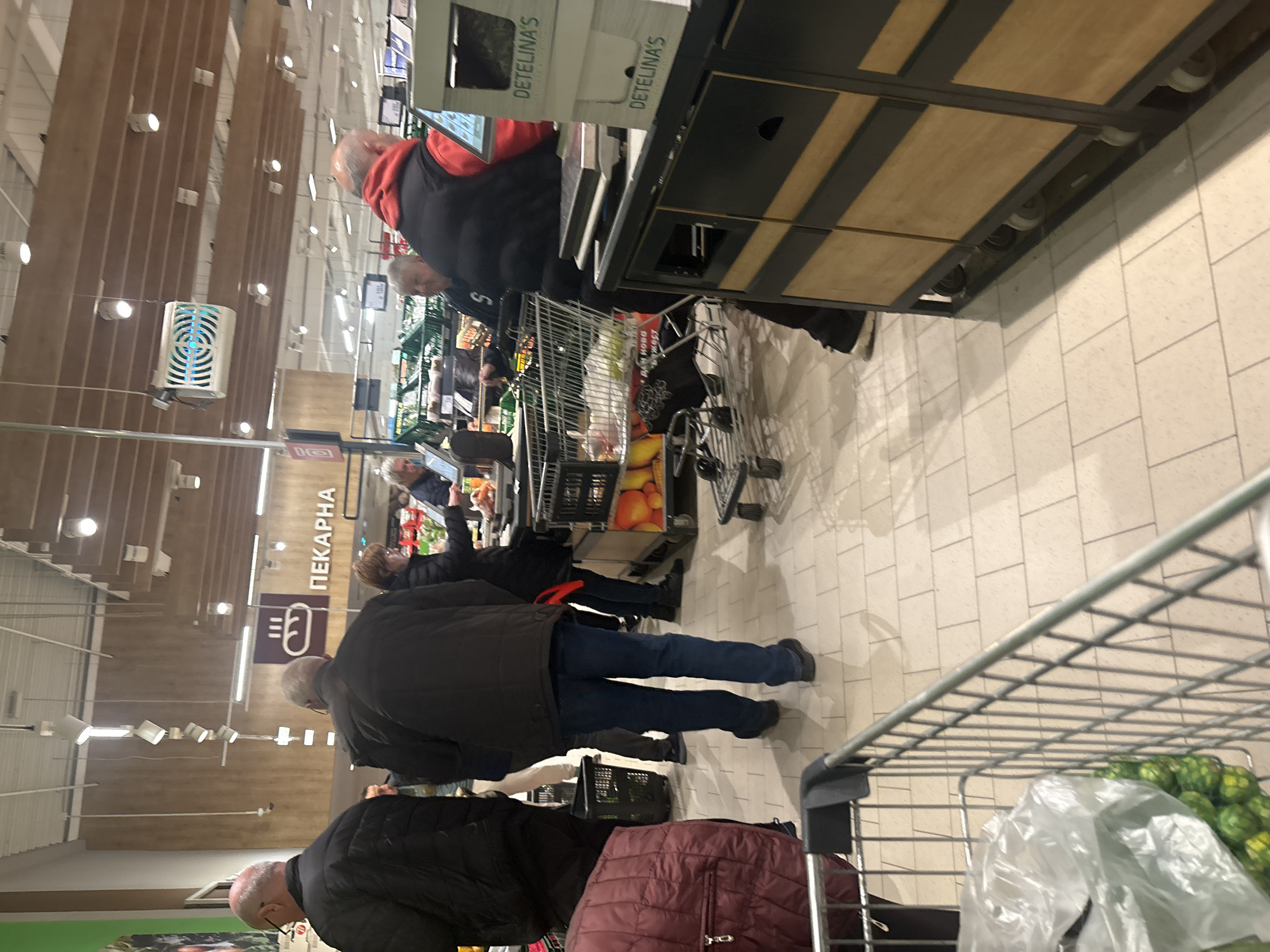 The image shows a checkout line in a store in Varna, Bulgaria. Customers with shopping carts are waiting to pay. Several people are wearing dark jackets. One person in line is wearing a red vest. The store appears to be a supermarket, based on the items visible in the shopping carts. There is a sign with Cyrillic text, including the word "ПЕКАРНА", likely indicating a bakery section.