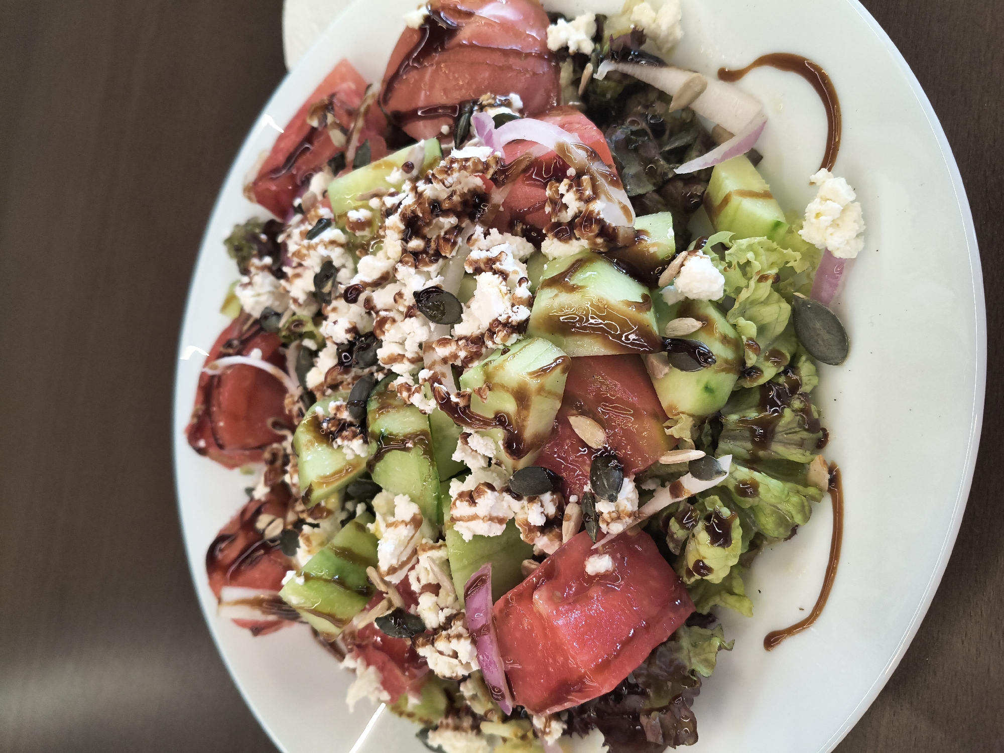 A close-up overhead shot features a vibrant watermelon and cucumber salad served on a white plate. The salad is generously topped with crumbled feta cheese, pumpkin seeds, and slivered almonds, all drizzled with a dark balsamic glaze. Visible ingredients include chunks of red watermelon, sliced green cucumber, purple onion slivers, and leafy green lettuce. The plate rests on a dark brown wooden surface, with a blurred white cloth visible on the right edge of the frame. The lighting is natural and bright, highlighting the fresh colors of the salad.