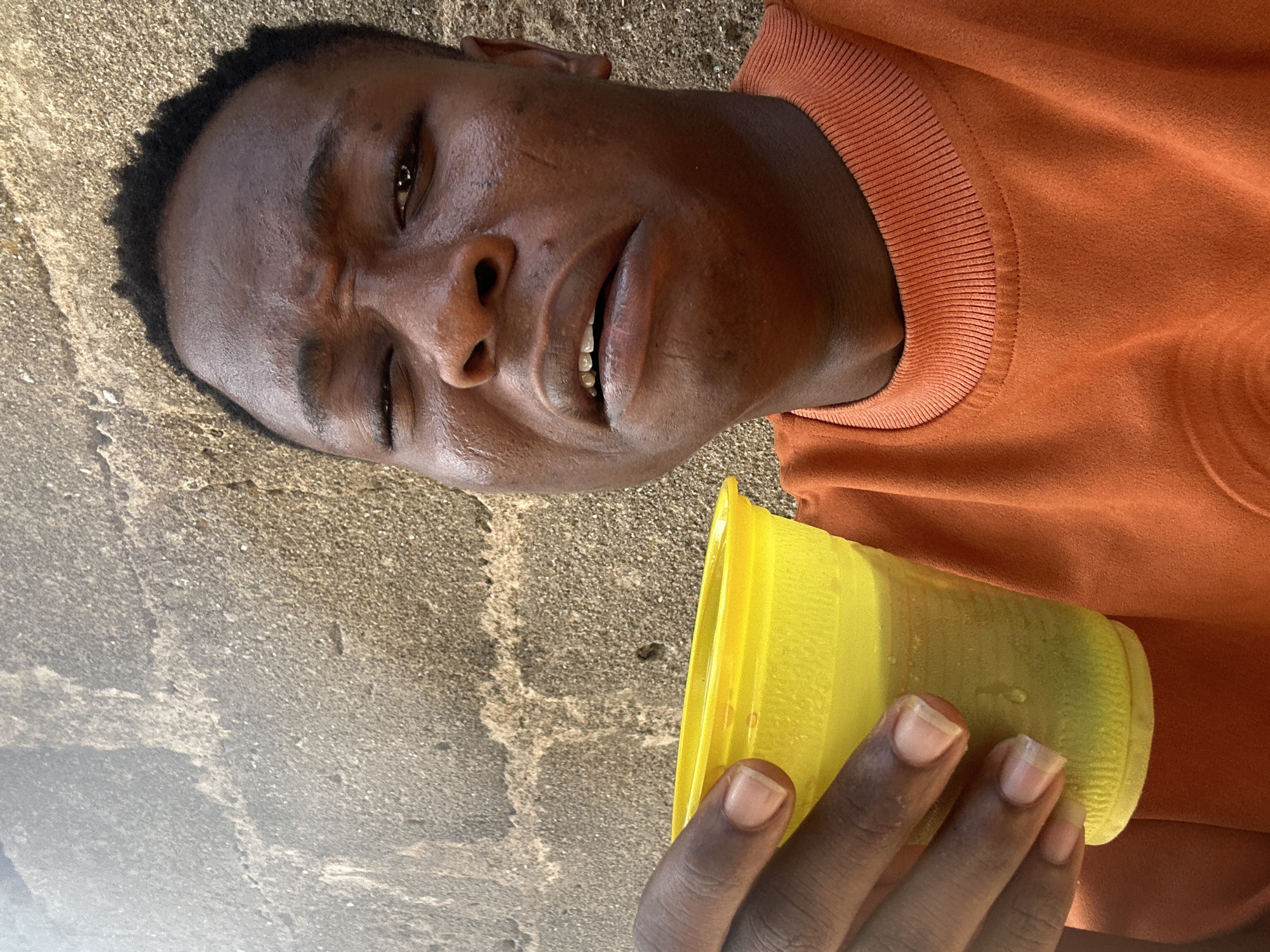 A dark-skinned individual with a slightly damp face, wearing an orange sweater, is lying on a concrete surface in the city of Unknown, Unknown. They are holding a yellow cup in their right hand. The setting appears to be outdoors, given the concrete floor.