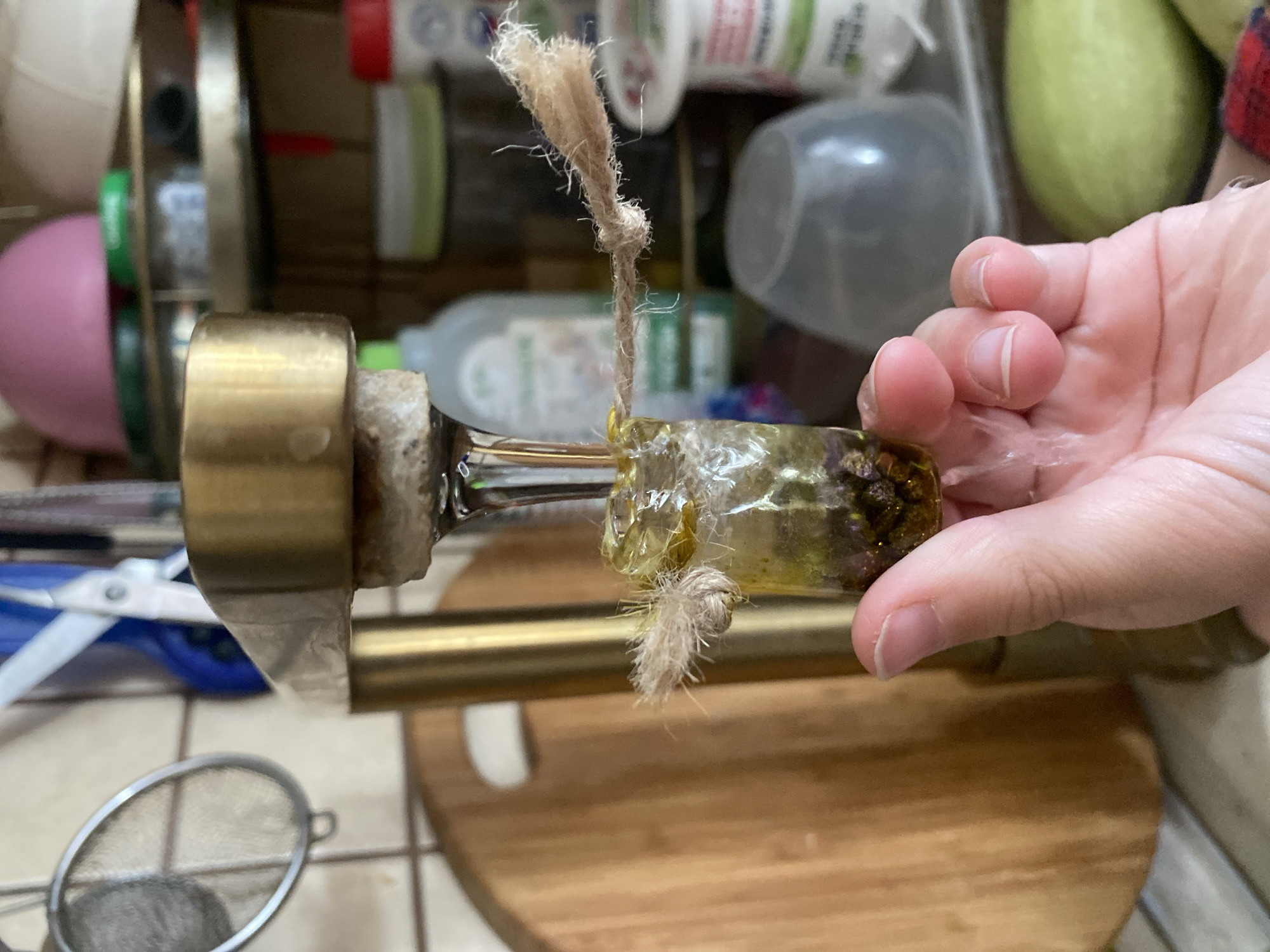 A person's hand is holding a small glass vial filled with a yellowish liquid and dark particulate matter. A piece of rough twine is tied around the neck of the vial, with frayed ends extending outwards. The vial is positioned beneath a brass-colored faucet, from which a thin stream of clear liquid is dripping into the vial. Some of the liquid has spilled onto the person's fingers. In the background, a wooden cutting board is visible, along with shelves containing various containers and objects. The scene appears to be in a kitchen. The location is not specified as Volzhsk, Russia.