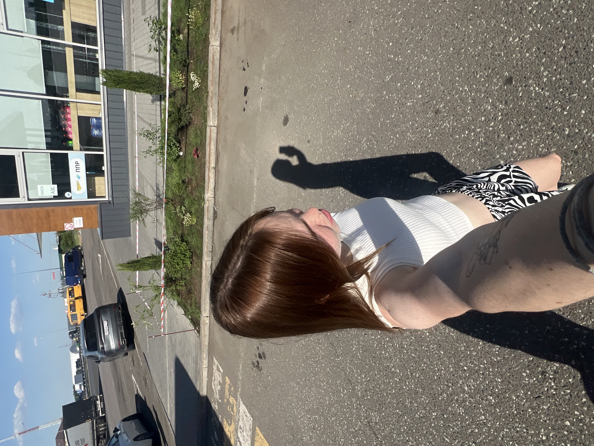 The image appears to be a selfie taken outdoors in Kazan, Russia. The perspective is from above, looking down. The subject is a person with reddish-brown hair wearing a white top and black-and-white patterned shorts. Their arm, which holds the camera, is visible in the lower right, showing a tattoo. The shadow of the person's hand is cast on the asphalt.
In the background, there is a building with large windows, landscaping including shrubs and flowers, and a parking area with several vehicles, including a grey car. Further back, there are buses. The sky is blue with some clouds.