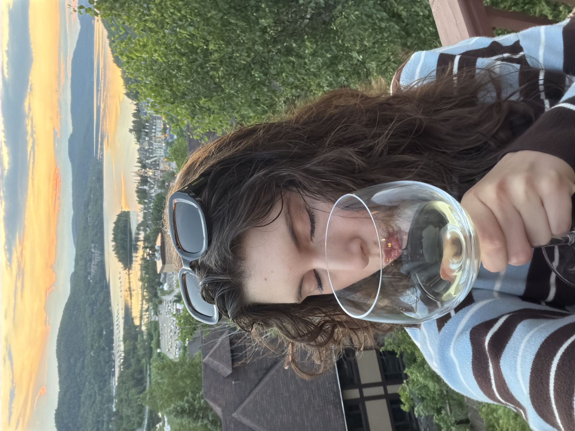 A young woman with long, curly brown hair and sunglasses perched on her head is holding a glass of white wine and drinking from it. She is wearing a blue and brown striped sweater. The background shows a scenic view of Mont-Tremblant, Quebec, Canada, with a lake, rolling hills covered in trees, and buildings in the distance, all bathed in the warm glow of sunset. The image appears to be taken from a balcony or deck, indicated by the presence of a wooden railing in the foreground.