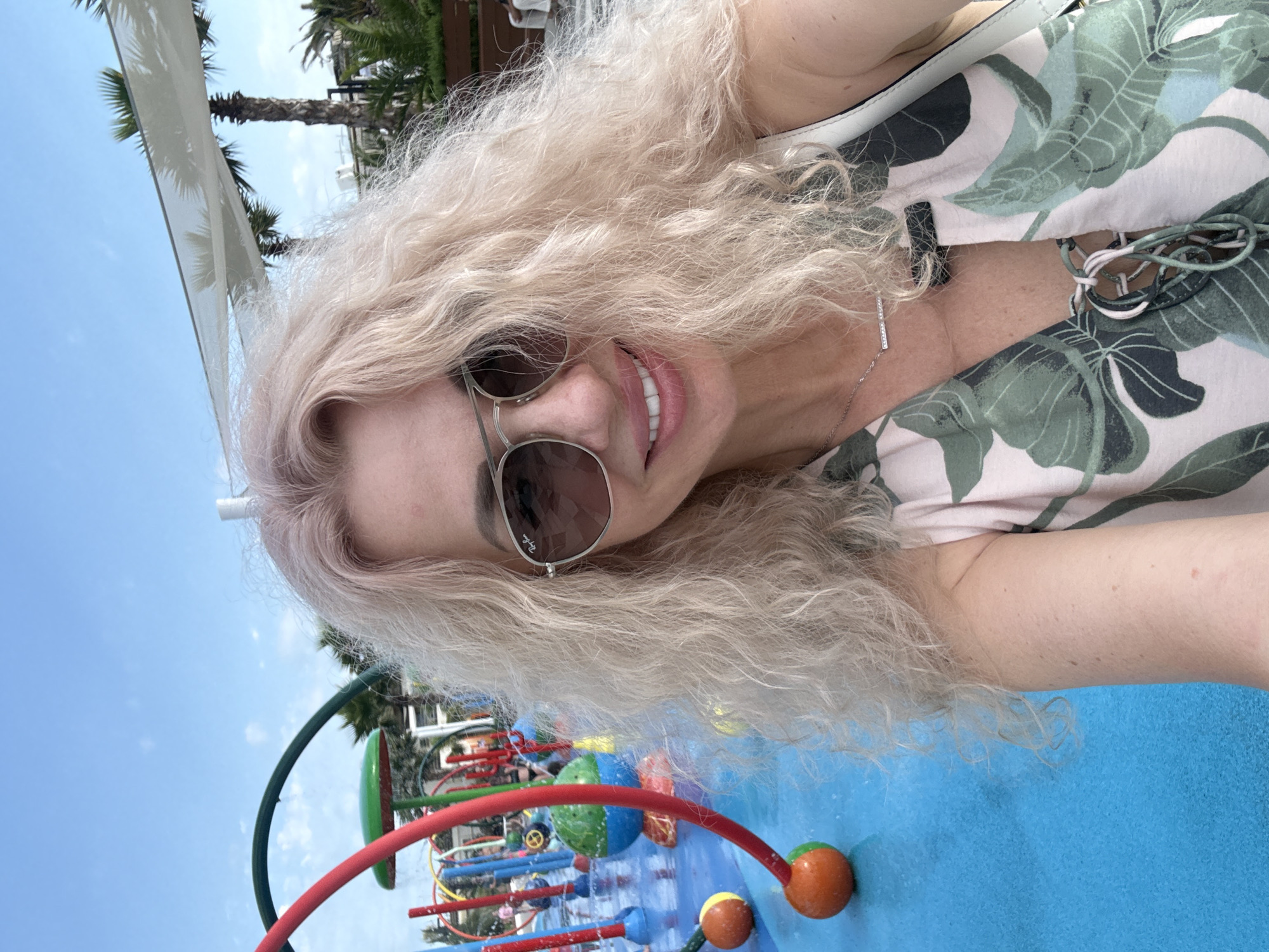 This is a close-up, selfie-style photograph of a woman with blonde, wavy hair and sunglasses, taken at an outdoor water park. She is smiling and wearing a floral-patterned top. In the background, there is a blue water play area with colorful splash pad features and palm trees, consistent with the environment of a resort in Sochi, Russia. The sky is clear and blue.