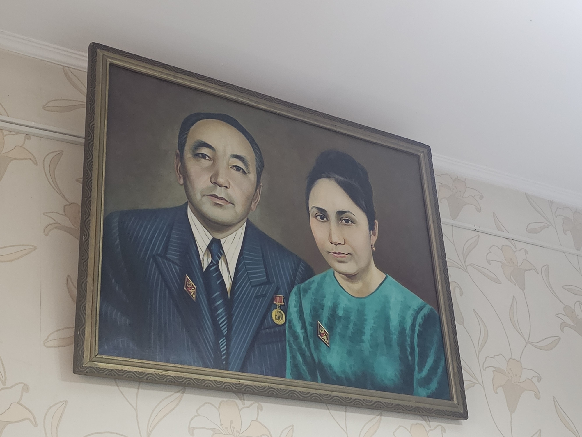 A framed portrait of a man and a woman hangs on a floral-patterned wallpaper. The man, on the left, is wearing a dark pinstripe suit with a white shirt and a blue tie. He has dark hair, dark eyes, and a serious expression. On his left lapel, he wears a red and gold medal. The woman, on the right, is wearing a teal-colored garment. She has dark hair pulled back and a neutral expression. She wears a small gold earring and a red and gold star-shaped pin on her chest. The portrait is lit from above and to the right, casting subtle shadows. The wallpaper has a light background with a repeating pattern of stylized lilies in pale yellow and cream. The ceiling, visible at the top of the image, is white and has a white crown molding. The overall impression is of a formal and possibly commemorative portrait. The location is identified as a residential interior in Ak-Jol, Kyrgyzstan.