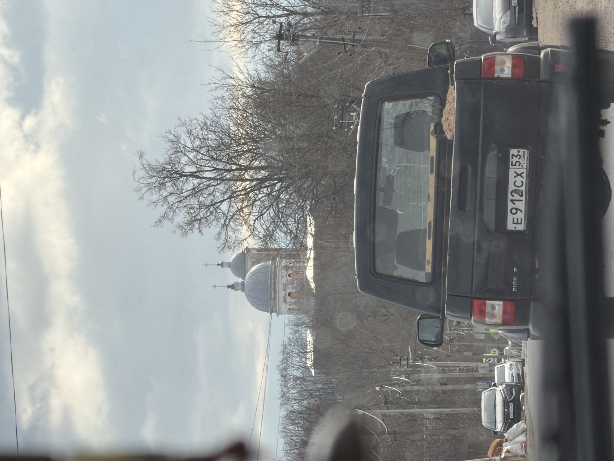 The image captures a street scene in Bronnitsy, Russia, as viewed from inside a vehicle. In the foreground, the rear of a dark-colored pickup truck is prominently displayed, with a Russian license plate reading "E912CX 53." A second car can be seen in front of the pickup truck. Beyond the trees and telephone poles, there are two domes of a church. The sky is overcast. The overall environment suggests a late fall or early spring season, based on the absence of leaves on the trees.