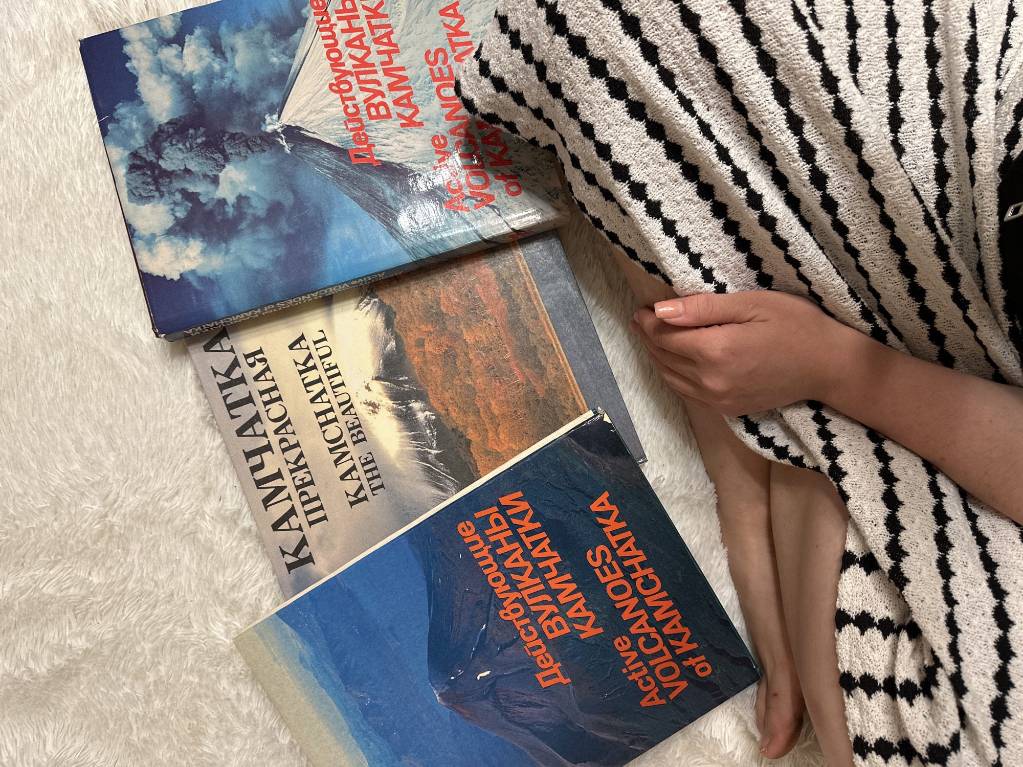 The image shows three books about Kamchatka, Russia, stacked on a white, fluffy surface, possibly a rug or blanket. The books are: "Active Volcanoes of Kamchatka" (both in English and Russian) and "Kamchatka the Beautiful" (also in both languages). A person is partially visible on the right side of the image, covered by a patterned blanket or sweater with alternating black and white stripes. Their hand rests on the blanket, and their leg is partially visible. The environment appears to be indoors. There is no information on this image to suggest it's in the city of Nizhny Novgorod.