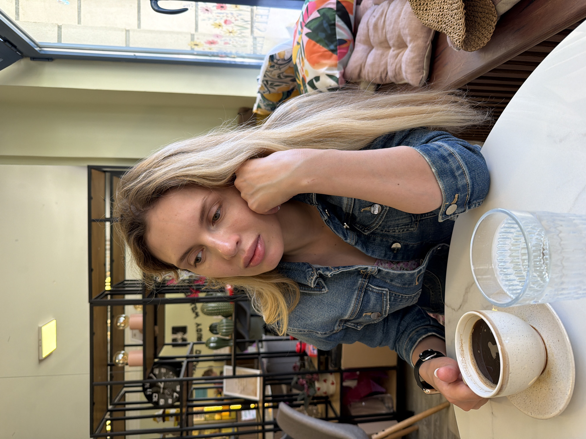 A woman with long, blonde hair, wearing a denim jacket, is sitting at a white table in what appears to be a café. She is holding her head with one hand and has a cup of coffee and a glass of water on the table in front of her. The background features a wall with shelves displaying various decorative objects. Based on the café's interior design and the woman's appearance, the location is likely in the city of Budva, Montenegro, which is known for its modern cafes.