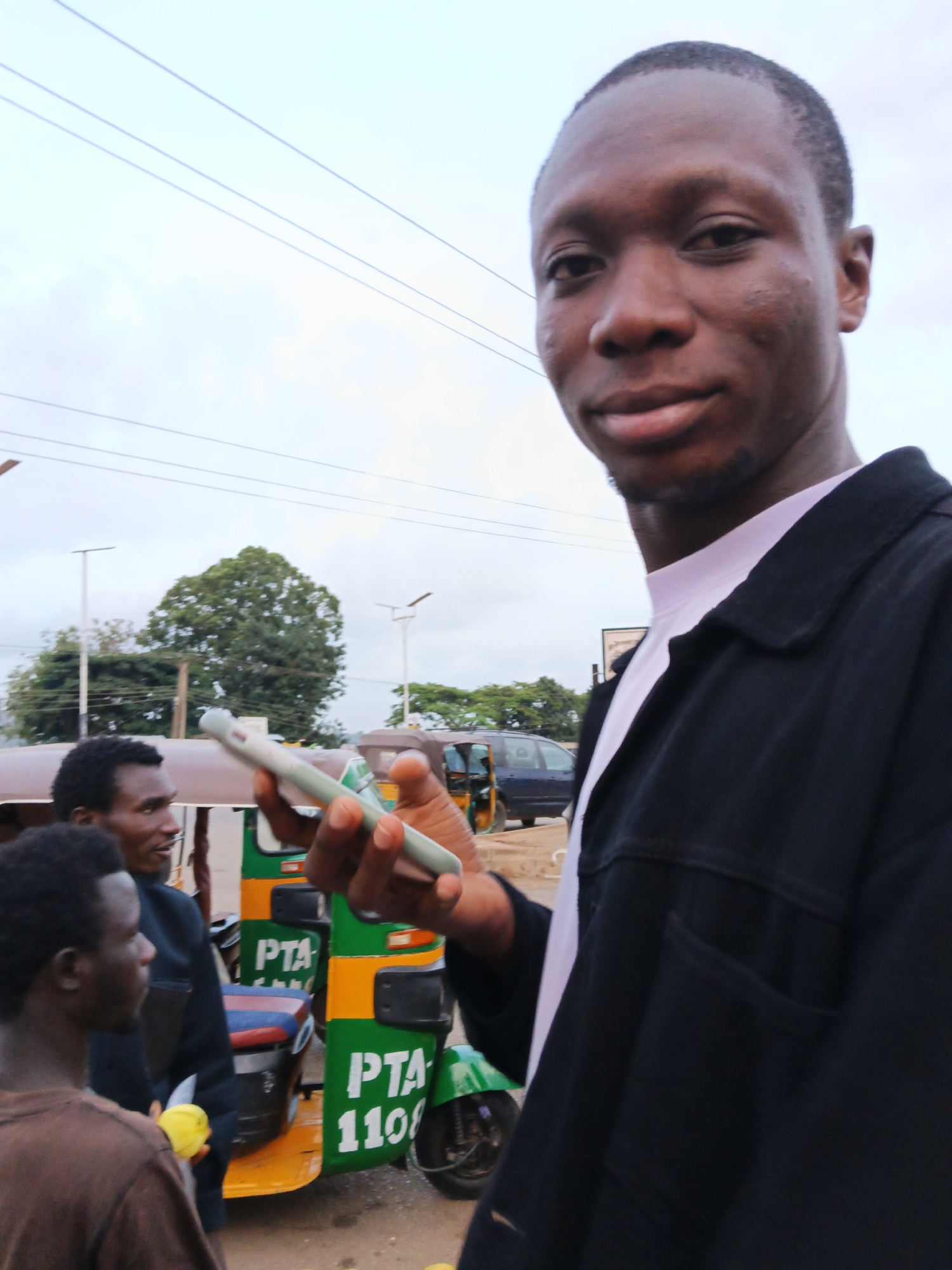 The image depicts a street scene with several people and vehicles. In the foreground, a young Black man wearing a dark jacket over a white shirt is holding a light-colored mobile phone in his right hand, looking towards the camera. To his left, another young Black man in a brown shirt is partially visible, looking to the left. Further in the background, a third Black man in a dark blue jacket is looking to the left, standing next to a green and yellow auto-rickshaw with "PTA" and "1108" written on it. Another auto-rickshaw and a dark blue van are visible behind them. The environment appears to be outdoors, with power lines visible in the sky, suggesting an urban or suburban setting. Trees and streetlights are also present in the background. The overall lighting suggests it is daytime, with an overcast sky.