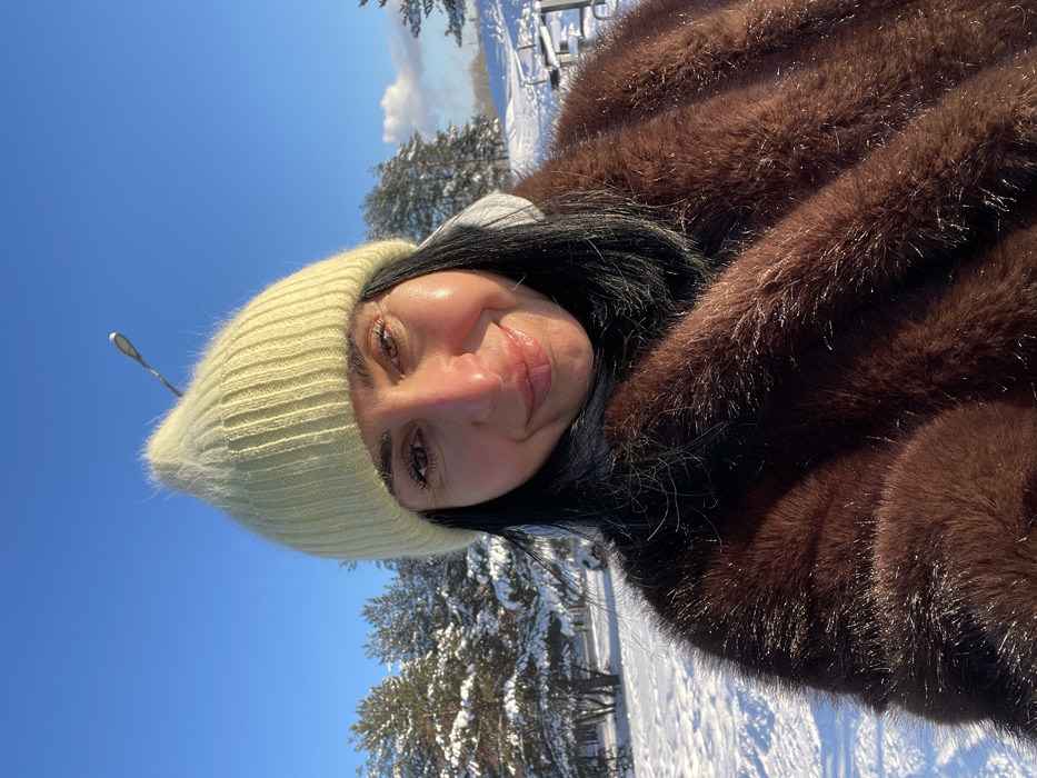 An outdoor portrait captures an adult female situated in a snowy environment within the city of Syasstroy, Russia, under a clear blue sky. The subject, positioned in the foreground, looks directly at the camera with a neutral expression and a slight smile. She wears a light yellow knitted beanie hat, a dark turtleneck or high-neck garment, and a brown fur coat, with a light grey hooded garment visible beneath the fur coat. Her dark hair frames her face, and her cheeks exhibit a rosy tone. The background reveals a landscape uniformly covered in white snow, with multiple snow-dusted evergreen trees. A black street light pole extends diagonally upwards from the upper center of the frame. On the far right, faint outlines of snow-covered outdoor seating structures, possibly benches or picnic tables, are discernible. A small, white cloud or vapor plume is visible on the distant horizon to the right. The scene is brightly illuminated by direct sunlight, indicating a clear winter day.