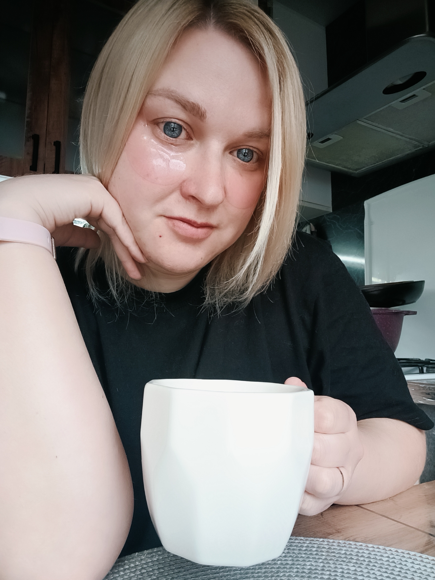 A blonde woman with blue eyes, possibly in Chaikovsky, Russia, is captured in a close-up indoor shot, likely a selfie. She has shoulder-length blonde hair, parted on the side, and a calm, direct gaze towards the camera with a subtle smile. Under-eye patches are visible on her cheeks beneath her eyes, suggesting a skincare routine. She wears a plain black short-sleeved top. On her left wrist is a light pink smart band or watch.
Her right hand holds a white, geometric-shaped mug, indicating she might be enjoying a warm beverage. A thin ring is visible on her right ring finger. She rests her left arm on a surface, with her hand supporting her chin.
The background reveals a home kitchen setting. To the left, dark wooden cabinets are partially visible. On the right, a grey range hood or extractor fan is positioned above a gas stovetop with a pan and a dark purple pot. A white refrigerator or cabinet door is also present on the far right. The mug rests on a grey textured placemat or table runner, which is placed on a wooden countertop. The lighting appears to be natural daylight, suggesting it is daytime, possibly morning or early afternoon. No visible text is present in the image.