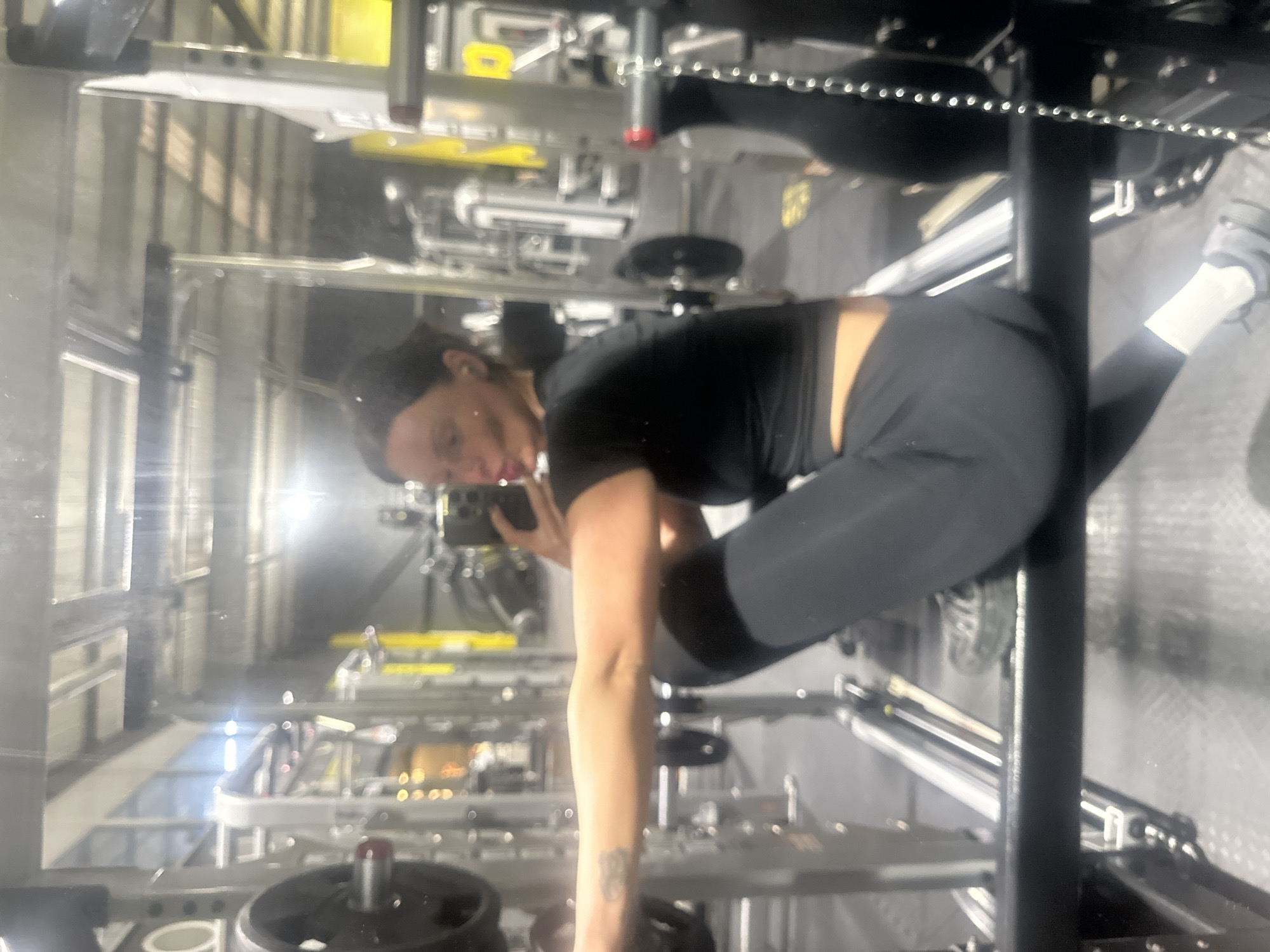 A woman in black workout clothing is seen in a mirror at a gym, likely taking a selfie with her phone. She is wearing black leggings, a black short-sleeved top, white socks and athletic shoes. Gym equipment is visible in the reflection and in the foreground. The location is likely in Tonbridge and Malling, United Kingdom.