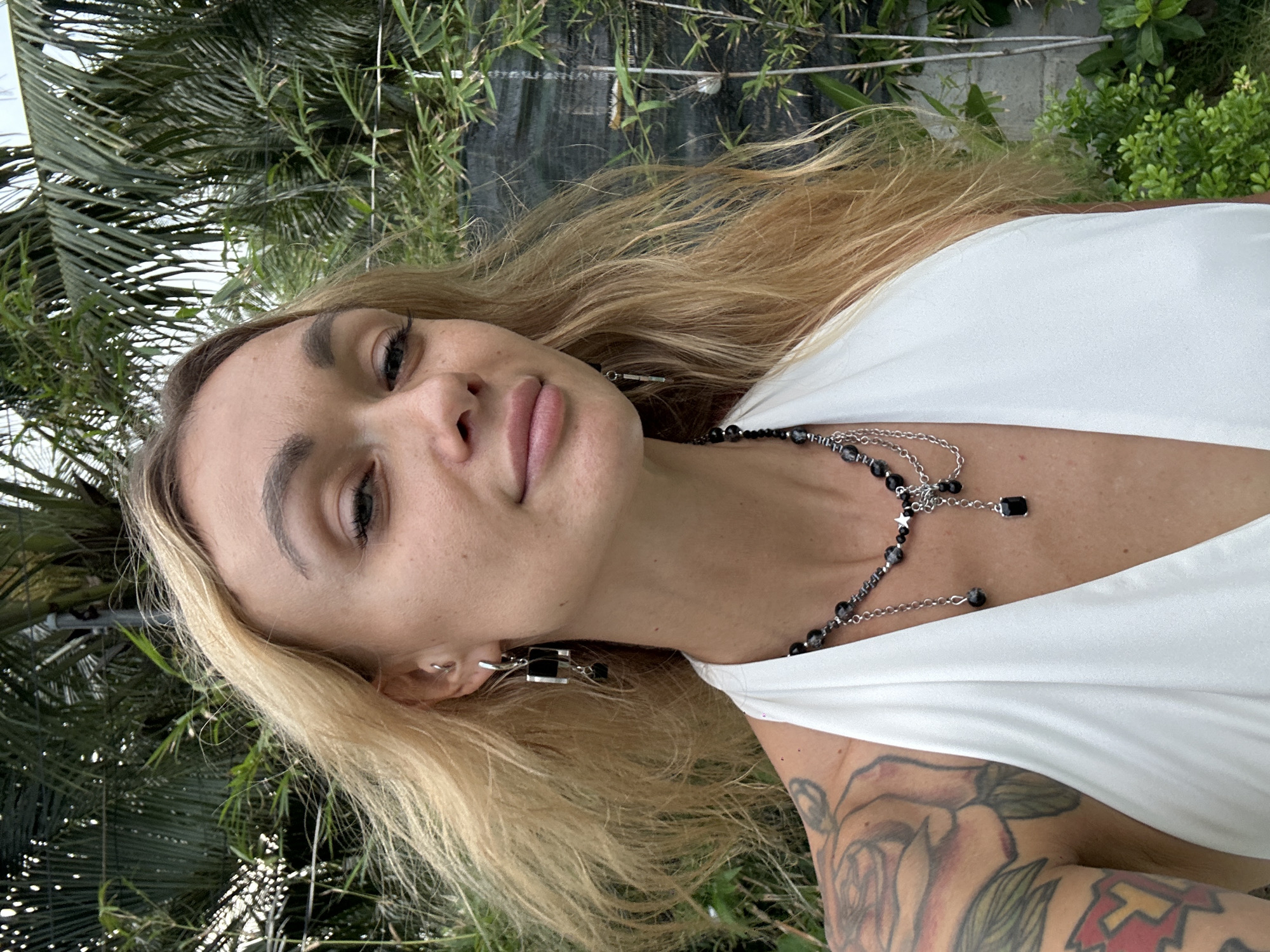 The image shows a woman with blonde hair and tattoos on her arm, wearing a white top and a black and silver necklace. She is outdoors, surrounded by greenery, possibly in a garden or park. The background includes various types of plants, including palm trees and bamboo. The location is unspecified in the prompt other than Mũi Né, Vietnam.