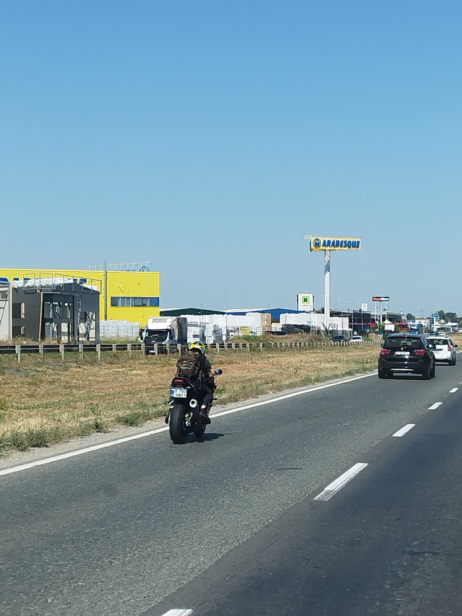 A motorcycle with a rider wearing a helmet and backpack travels on a highway. The rider is in the foreground, with the motorcycle's rear visible. In the background, there are several vehicles, including cars and trucks, on the highway. There are also buildings and a large yellow structure with the word "ARABESQUE" on a billboard. The environment appears to be an industrial or commercial area on the outskirts of a city. The location is identified as being in Chitila, Romania. The weather is clear and sunny with a blue sky.