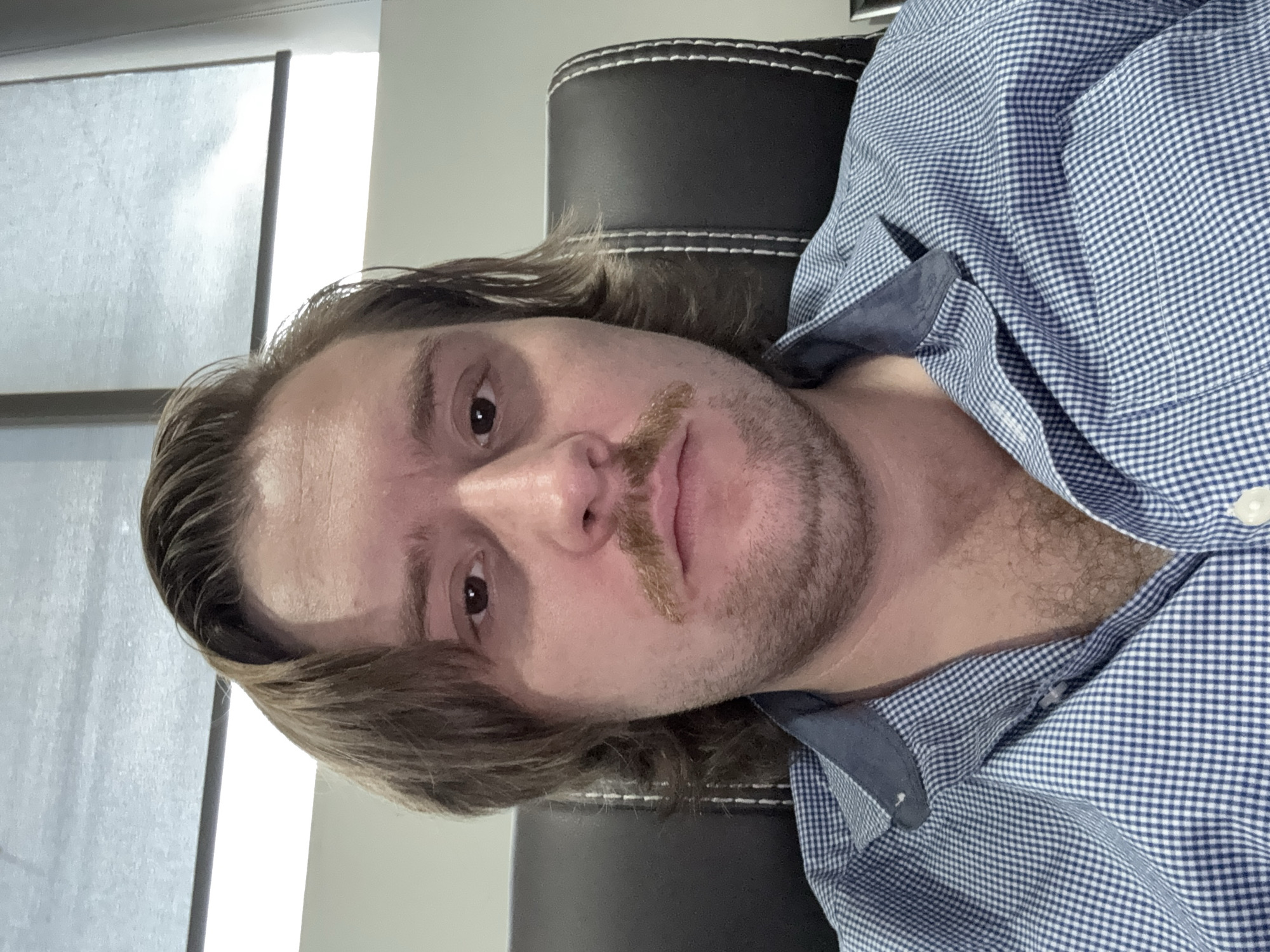 A selfie of a fair-skinned man with brown hair and a mustache. He is wearing a blue and white checkered shirt and sitting in a black leather chair. The background is a blurry light gray wall with a window. There is no information to suggest the image was taken in Cuautitlán Izcalli, Mexico.
