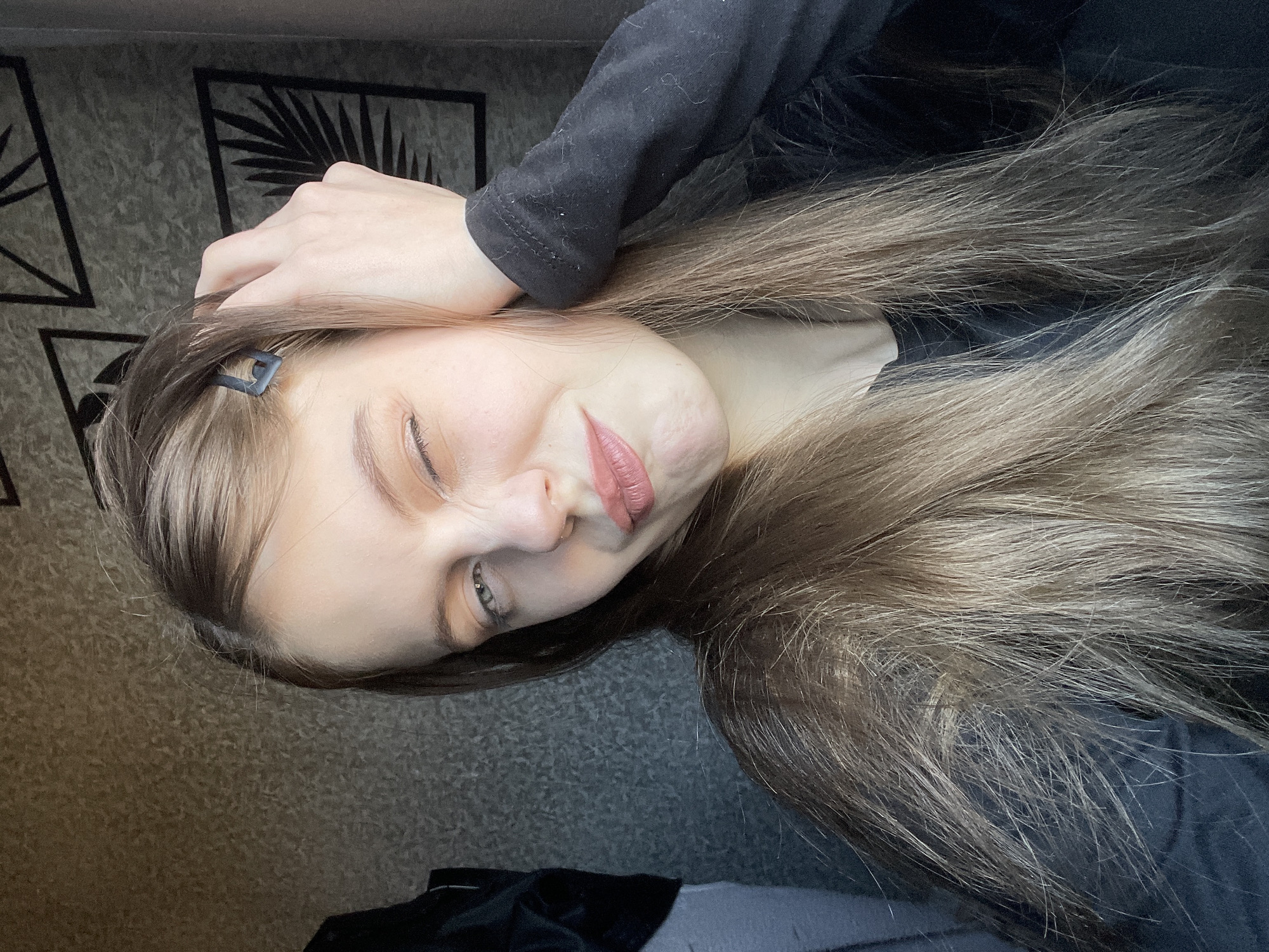 A young woman is lying down, her head resting on her hand. She has long, light brown hair with some blonde highlights, and a small, black hair clip is visible on the left side of her head. She is wearing a black long-sleeved shirt. One eye is closed, and she appears to be winking or squinting. The background consists of a textured gray wall. There is no evidence of her being in Фоки, Russia.