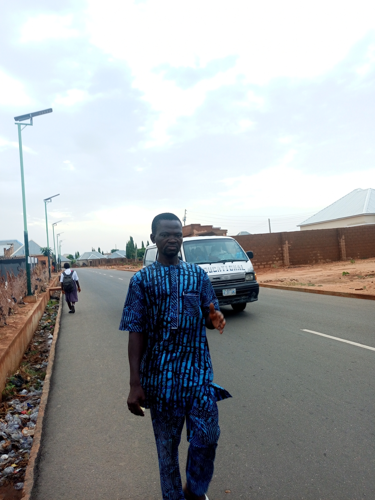 A man wearing a blue and black patterned shirt and pants is walking on a gray asphalt road. A white van with "EDUCATIONAL" written on it is behind him. A person in a school uniform is walking on the left side of the road. Tall street lights are visible along the road. Debris is present in the gutter on the left side of the road. A brown brick wall lines the right side of the road. The sky is cloudy. The location is a street in what appears to be the city of Tempure, Nigeria.