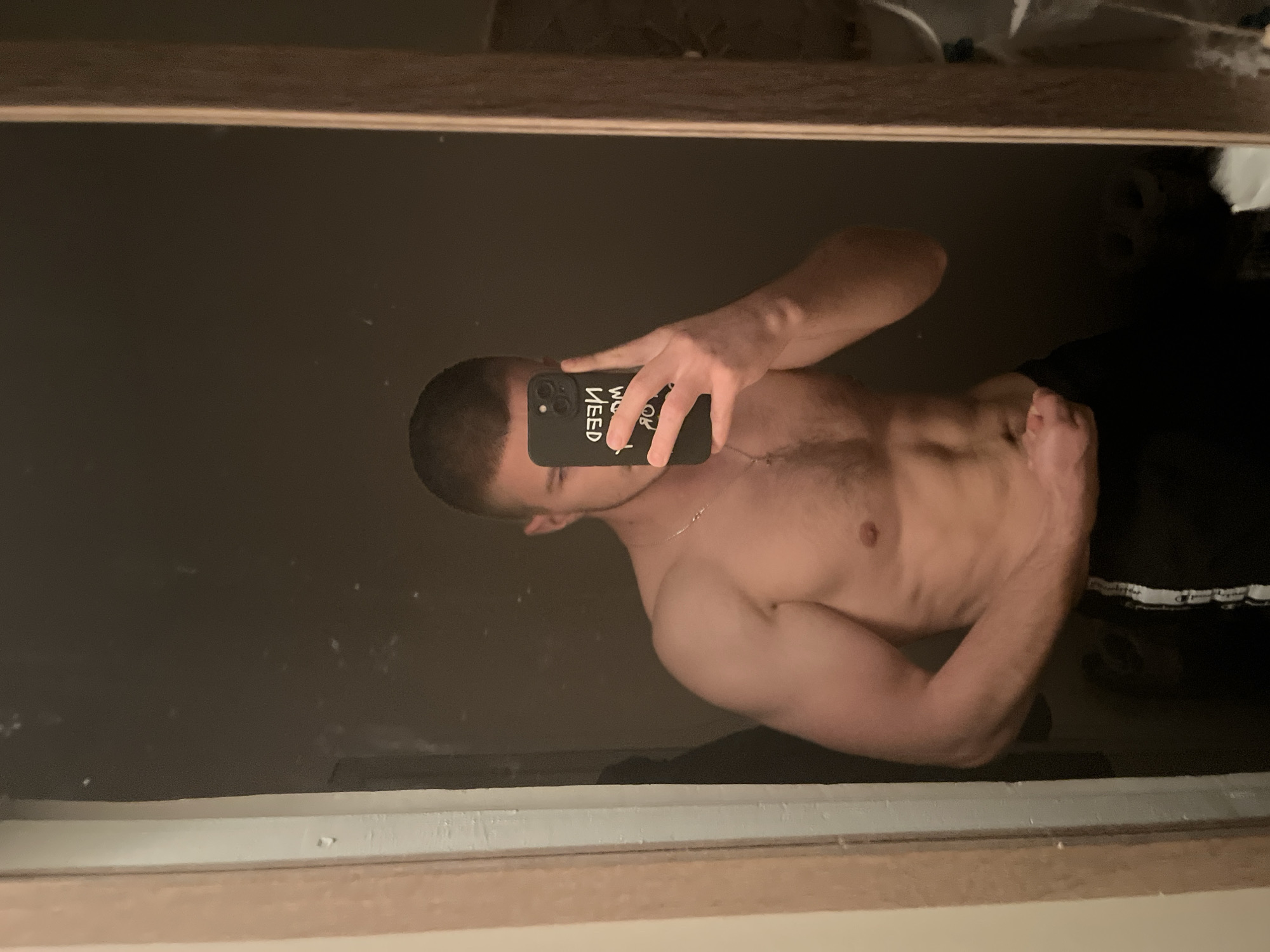 A shirtless man is taking a selfie in a mirror. He is holding a black iPhone with a case that has white text. The man has short, dark hair and a necklace. He is wearing dark pants. The environment appears to be a dimly lit room. The location is unknown.