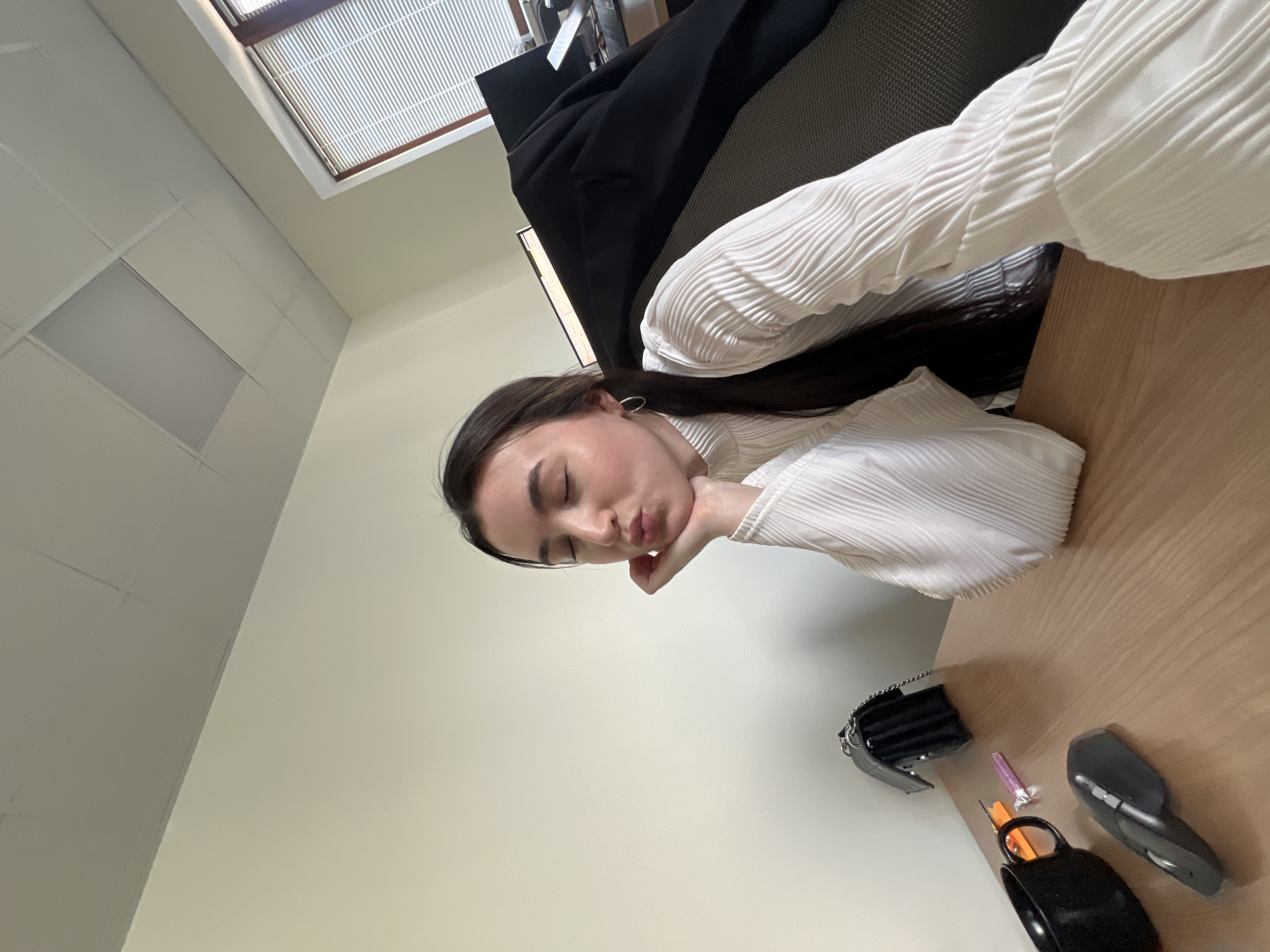 A young woman with dark hair is sitting at a wooden desk in what appears to be an office. She is wearing a white, long-sleeved shirt with a textured fabric. Her eyes are closed, and her chin is resting on her hand, with her lips puckered as if she is blowing a kiss. On the desk, there is a black mug, a computer mouse, a black handbag, and a couple of small items that are difficult to identify. The room has plain, light-colored walls and a drop ceiling with fluorescent lighting. A window with blinds is visible in the background, as well as a dark jacket draped over a chair. The location is not specified as Kazan, Russia.