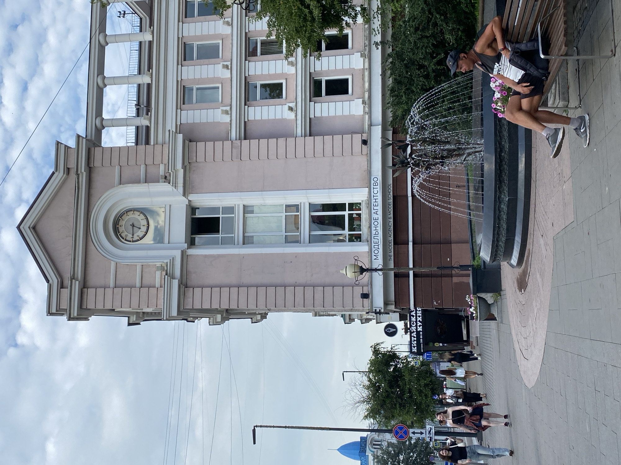This image depicts a street scene in Krasnoyarsk, Russia, featuring a building with a prominent clock and a fountain. The building, painted in a light pink hue, has classical architectural elements including columns and arched windows. A sign on the building reads "МОДЕЛЬНОЕ АГЕНТСТВО" and "THE MODEL AGENCY & MODEL SCHOOL" in English. In the foreground, a fountain sprays water, with a man sitting on a wooden bench to its right. He is wearing a black baseball cap, a grey tank top with white lettering, black shorts, and grey athletic shoes, with his legs crossed and one arm resting on his lap. He appears to be looking towards the fountain. To the left of the building, several women are walking on a sidewalk. One woman in the front wears a black top and blue jeans, while another behind her has a black strapless top, a blue skirt, and a red shirt tied around her waist. Further down the sidewalk, other pedestrians are visible. Across the street, a building with a blue dome is partially visible, and a street lamp stands tall. The sky is overcast with clouds.