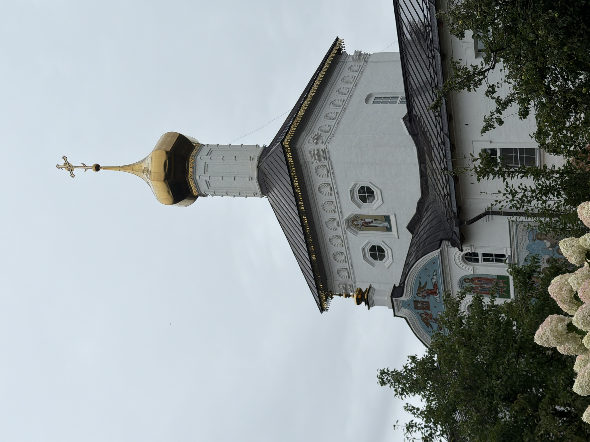 The image shows a white church with a golden dome and cross against a cloudy sky. The church has a dark roof and decorative details along the eaves. There are also religious murals visible on the exterior of the building. Trees and flowering bushes are in the foreground, partially obscuring the lower part of the church. The scene is set in Rusanovo, Russia.