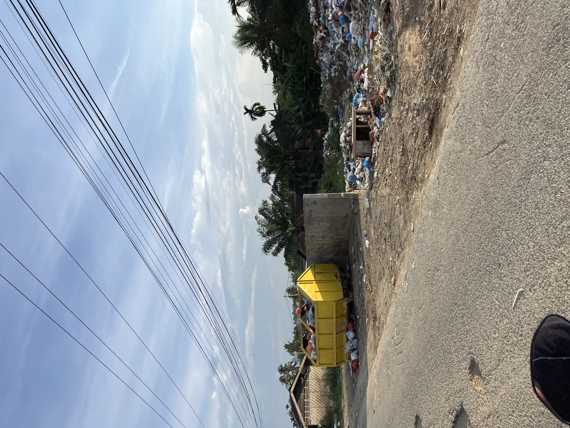 A bright yellow dumpster is overflowing with garbage. To the right of the dumpster, a large pile of trash, including plastic bags and debris, is scattered across the ground. In the background, palm trees and other vegetation are visible, along with some buildings and a gray concrete wall. Overhead, multiple electrical wires are strung across the sky, which is partly cloudy. The scene appears to be a roadside dump site in Alurcucur, Indonesia.