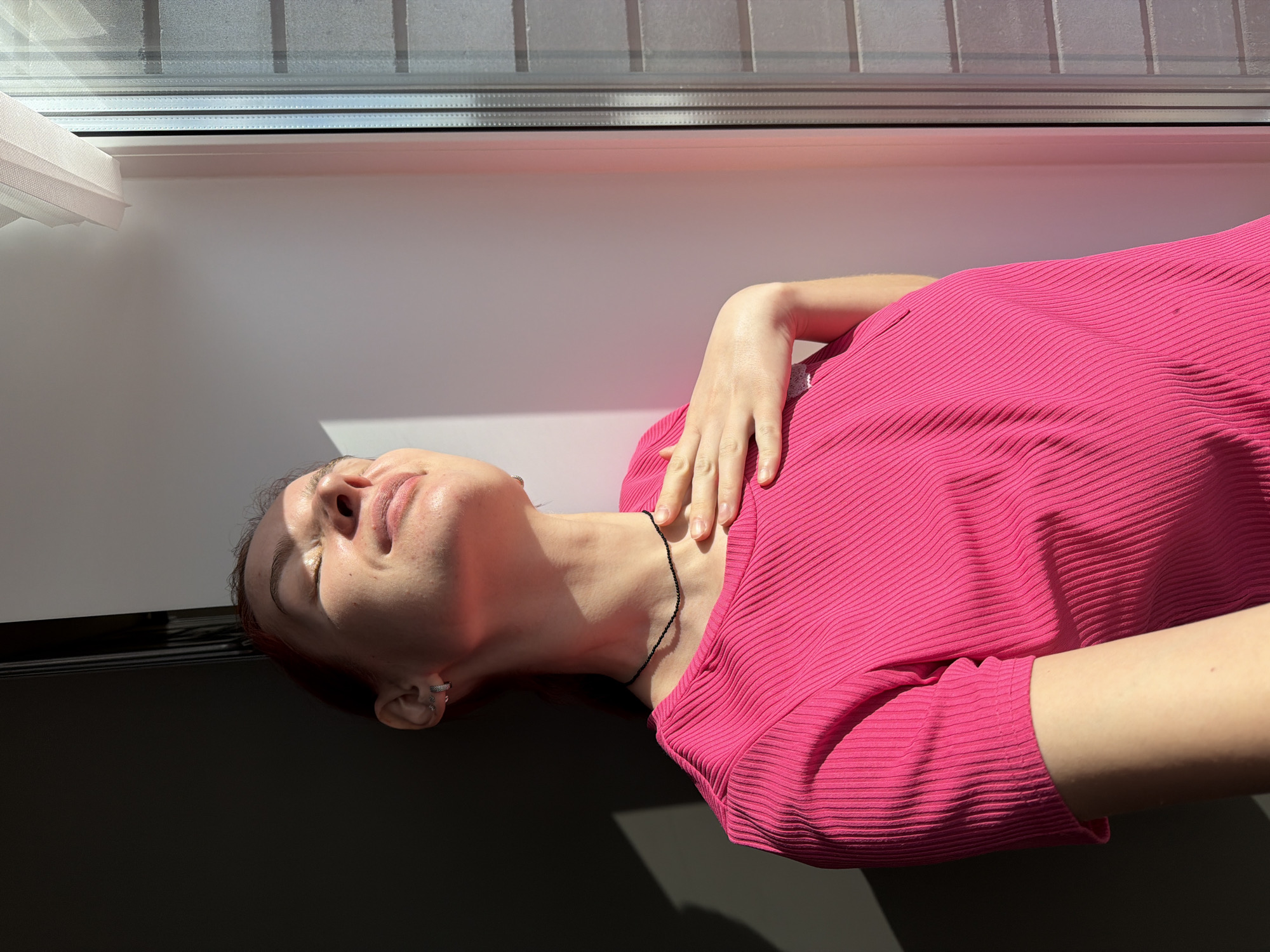 A woman is lying down indoors near a window. The scene is brightly lit by sunlight. The woman is fair-skinned and wearing a bright pink, ribbed top. She has a delicate black necklace around her neck and small, circular earrings. Her head is tilted back. Her right hand is placed gently on her collarbone. The background includes a window with visible horizontal window blinds and a white window frame. The lower left portion of the image is a dark gray or black surface. There is no information to determine the location in Chaikovsky, Russia.