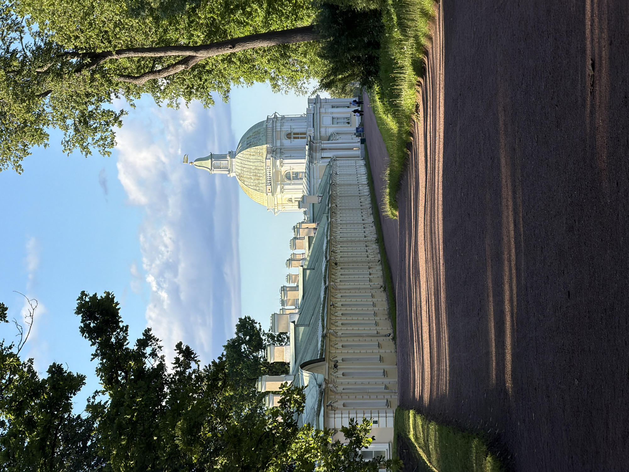 A pathway of dark gravel curves through a park. On the left, a long, pale yellow building with a green roof and numerous chimneys stretches into the distance. On the right, a large tree with green foliage frames the scene. In the background, a large white building with a green dome topped by a small tower and flag is visible. The sky is blue with scattered white clouds. Sunlight casts long shadows across the pathway from the trees. Two people are walking on the pathway near the buildings. The location is Unknown, Unknown.