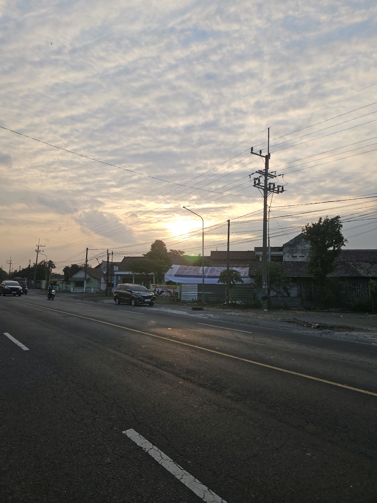 The image depicts a road scene in the city of Bandar Kedung Mulyo, Indonesia, during what appears to be sunrise or sunset. A paved road runs diagonally from the lower left to the upper right of the frame, marked with white and yellow lines. Several vehicles are visible on the road, including a black SUV, a motorcycle, and a white car. On the right side of the road, a cluster of buildings with tiled roofs are present, interspersed with trees. Utility poles with electrical wires crisscross the sky, which is filled with textured clouds and the warm glow of the sun. The overall impression is that of a rural or suburban road on the outskirts of a town.