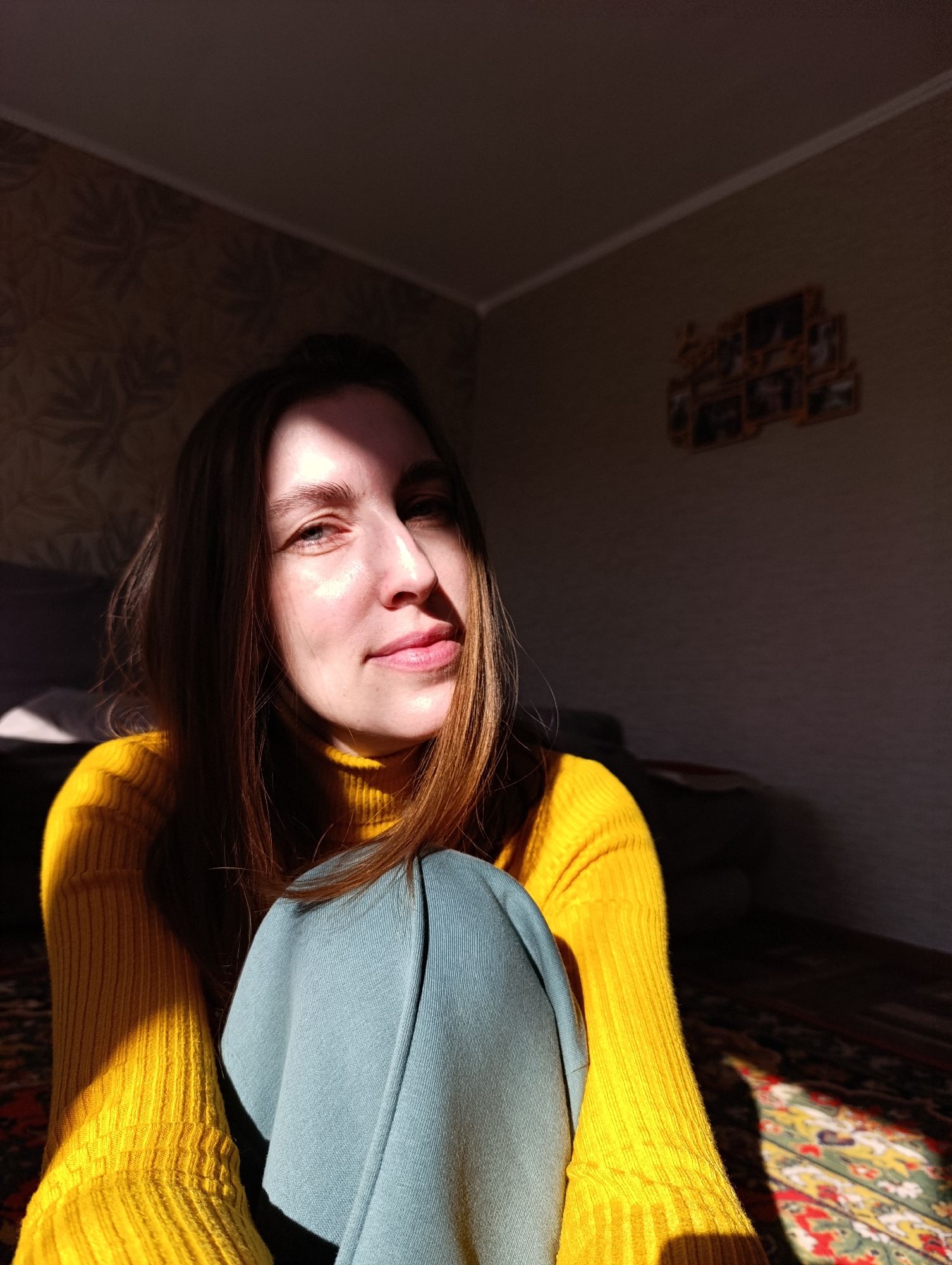 A woman is seated on the floor of a room. She wears a yellow turtleneck sweater and blue-green pants. Light illuminates her face and the front of her sweater, casting the rest of the room into shadow. Behind her is a sofa. On the wall is floral patterned wallpaper. In the upper right, a multi-picture frame hangs on the wall. The floor is covered with a patterned carpet. The location Фоки, Russia cannot be confirmed based on the visual information provided.