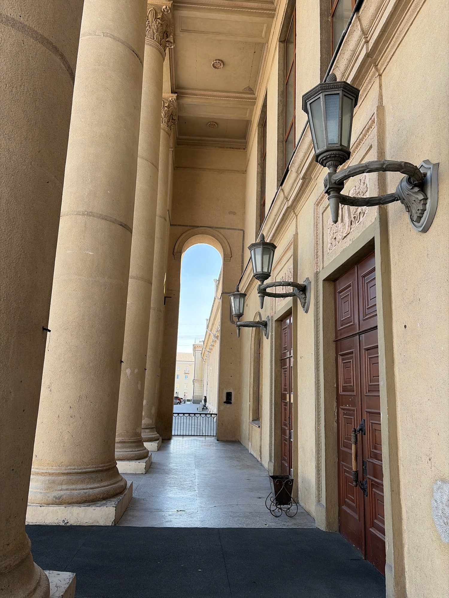 The image depicts a neoclassical colonnade with large, beige stone columns on the left side. A walkway runs alongside the colonnade, leading towards an arched opening in the distance. On the right side of the walkway, the facade of a building is visible, featuring a textured yellow wall, brown double doors with ornate handles, and several wall-mounted lanterns. Two of these lanterns are clearly visible, each with a hexagonal glass shade and an elaborate metal arm. Decorative relief work is present on the facade above the lanterns and around the doors. In the distance, through the archway, a street scene can be observed with buildings, a black metal fence, and a few parked cars. A small, decorative metal trash bin with wheels is situated near the walkway. The overall impression is one of historical architecture and an urban setting.