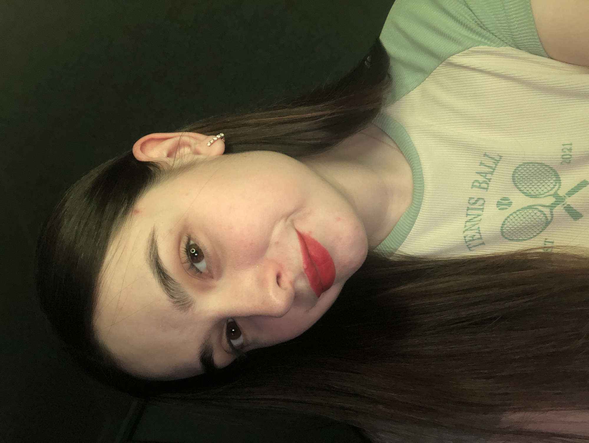 A person is facing the camera and looking down. They have brown hair, light skin, and are wearing red lipstick and a sparkly earring. They are wearing a light-colored shirt with green trim and a tennis ball design. The background is dark and out of focus. The location is not possible to determine from the provided image.