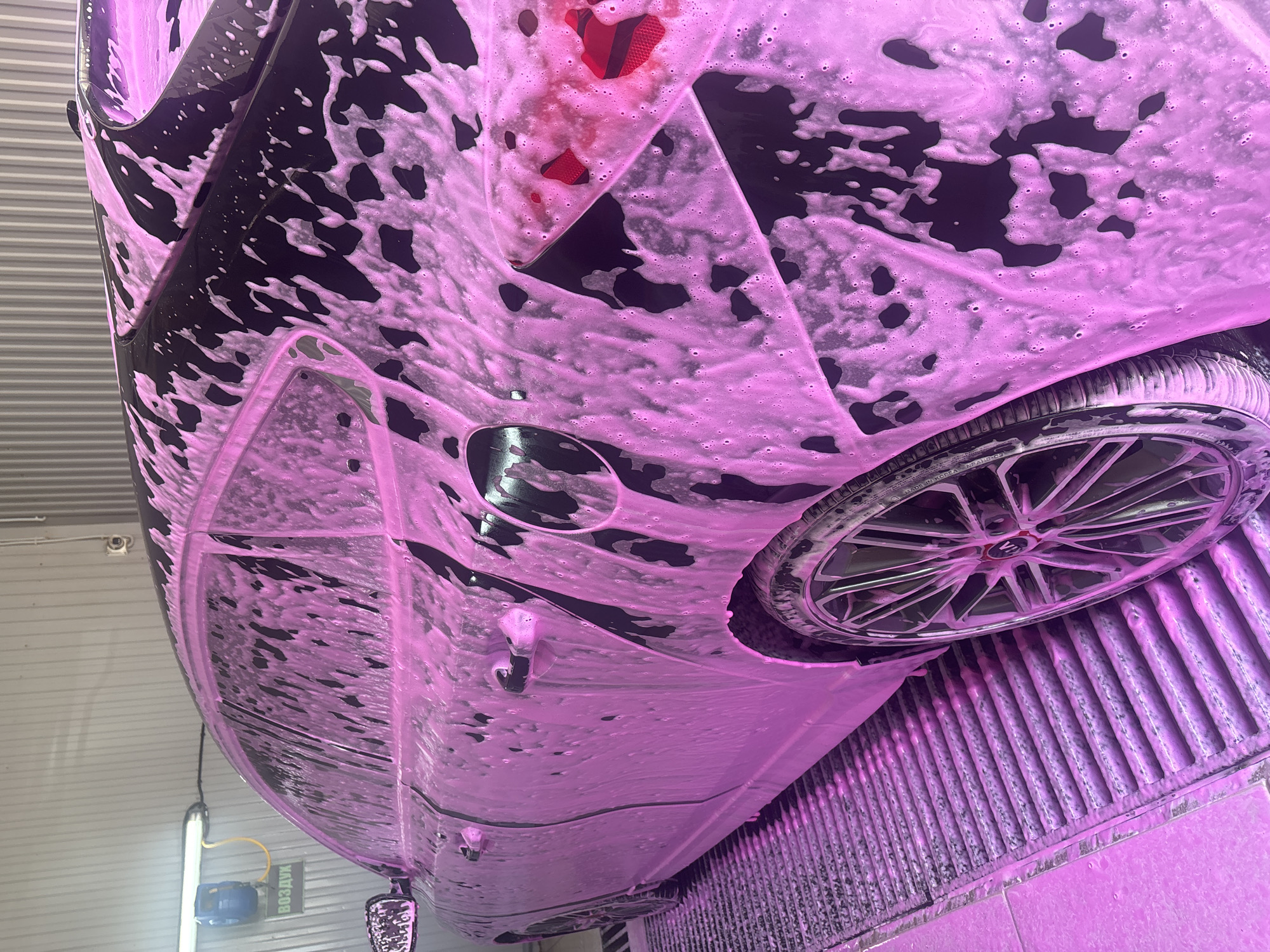 A low-angle, close-up shot depicts a black car heavily coated in pink foam, likely soap suds from a car wash. The rear of the vehicle is visible, including a taillight and a wheel with a multi-spoke design. The car is positioned inside what appears to be a car wash bay, as evidenced by the corrugated metal wall in the background and the grated floor, also covered in pink foam. A hose and signage are visible in the bottom left corner, though partially obscured. The location is Chaikovsky, Russia.