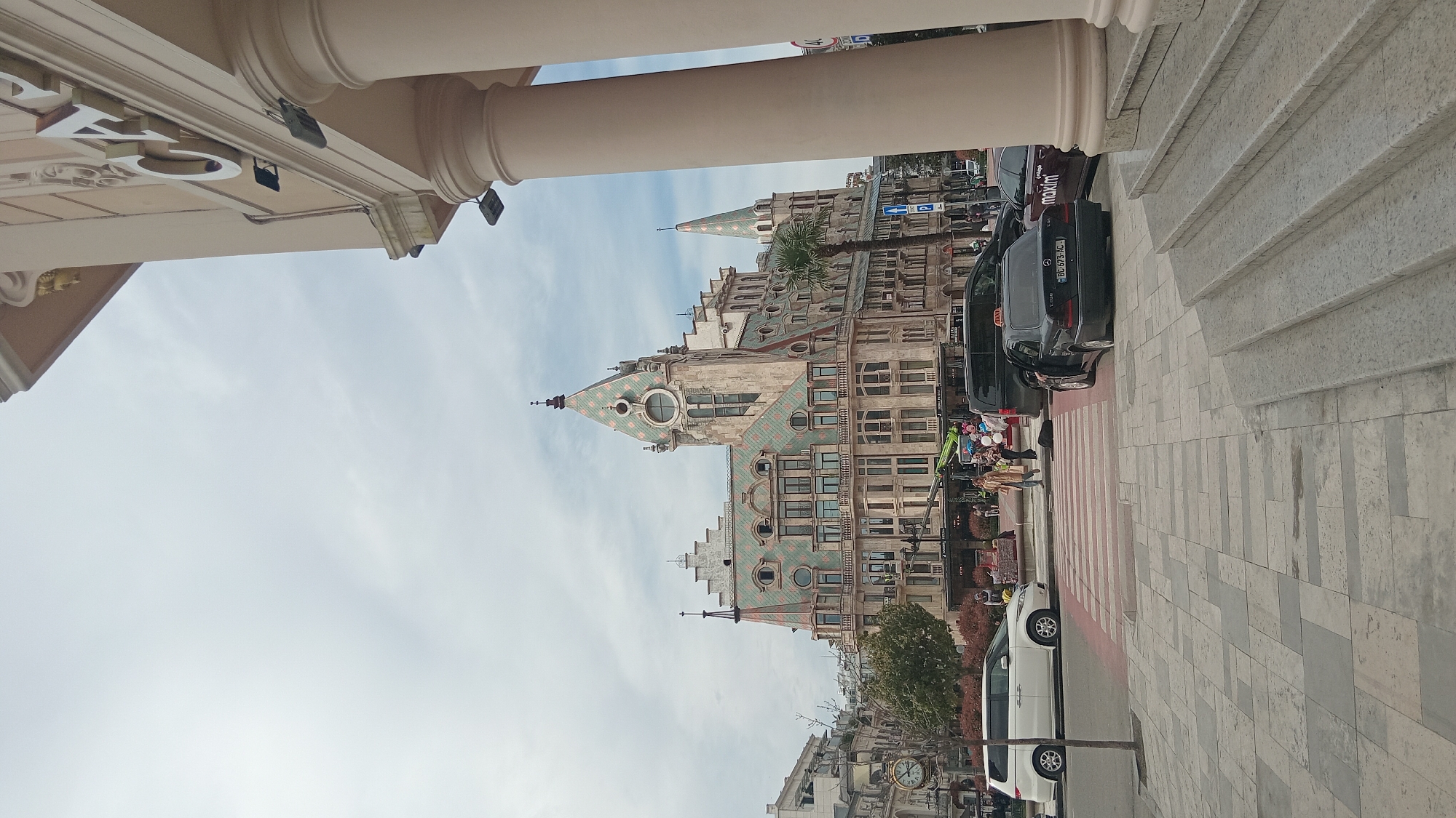 The image depicts a street scene in Batumi, Georgia. A large, ornate building with distinctive green and orange tiled roofs and multiple spires dominates the background. Several cars are parked along the side of the street, including a dark-colored taxi with "Maxim" branding. Pedestrians can be seen near a crosswalk. The foreground shows architectural elements, including columns and stairs, suggesting the perspective is from under a building or portico. The sky is overcast with a few wisps of clouds. The scene exhibits the unique architectural style characteristic of Batumi.
