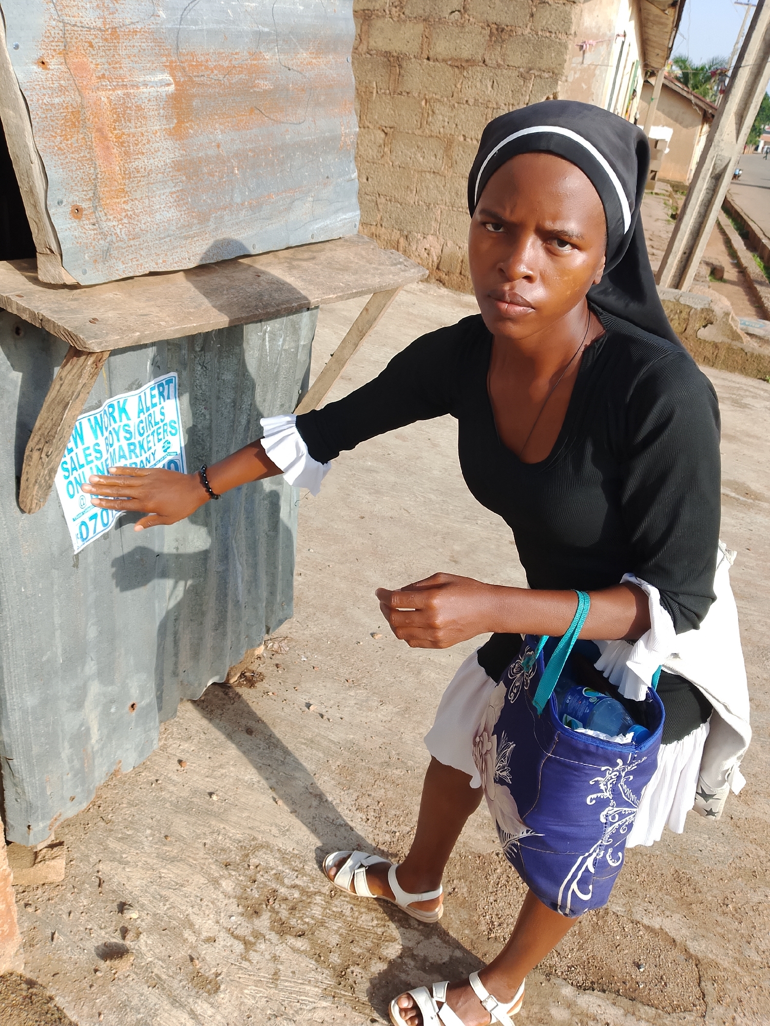 A young Black woman, wearing a black headscarf with white trim and a black long-sleeved top with ruffled white cuffs, and a white skirt, is standing in front of a corrugated metal structure with a wooden shelf attached. She has a blue bag with a white floral pattern slung over her shoulder and is wearing white sandals. Her right hand is extended, touching a flyer on the metal surface. The flyer is blue and white and contains text that includes "NEW WORK ALERT SALES BOYS/GIRLS ONLINE MARKETERS" and a phone number beginning with "070". The scene appears to be outdoors in an urban or semi-urban environment. The background shows a brick wall, wooden poles, and a dirt or paved surface. This setting is consistent with the environment of the city of Jos, Nigeria.