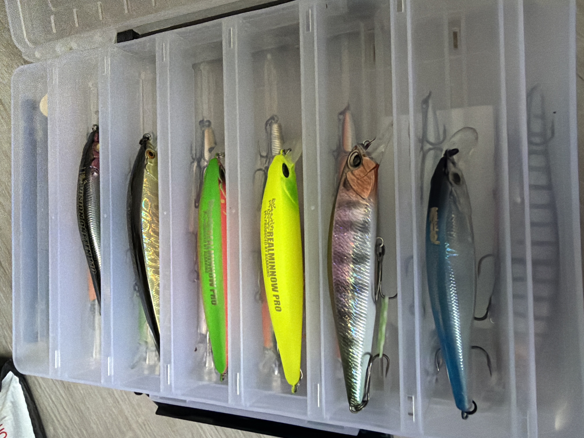 A tackle box is open and lying on its back on a light-colored wooden floor. The box is clear plastic and has multiple compartments, each containing a fishing lure. From top to bottom, the lures are: a silver and black lure with pink accents, a gold and black lure, a bright green lure, a bright yellow lure, a silver lure with iridescent purple and pink scales, and a blue and silver lure. All of the lures have hooks. The tackle box is open to the right, revealing its contents. The background is out of focus, but appears to be a light-colored surface.