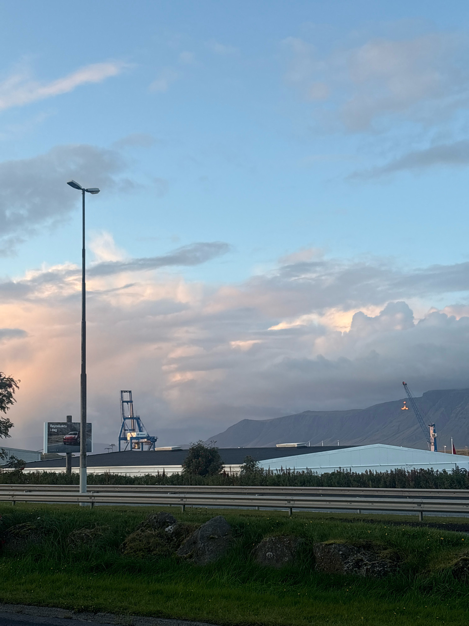 This image shows a view of Reykjavik, Iceland, looking towards the coast and mountains. In the foreground, there is a grassy area with some rocks and a street light. In the mid-ground, there is a highway with metal barriers. Beyond the highway, there are several industrial buildings and a large blue crane. In the background, there are mountains. The sky is blue with some clouds, and the sun appears to be setting, casting a warm glow on the clouds. On a billboard, a red car is advertised with Icelandic text.