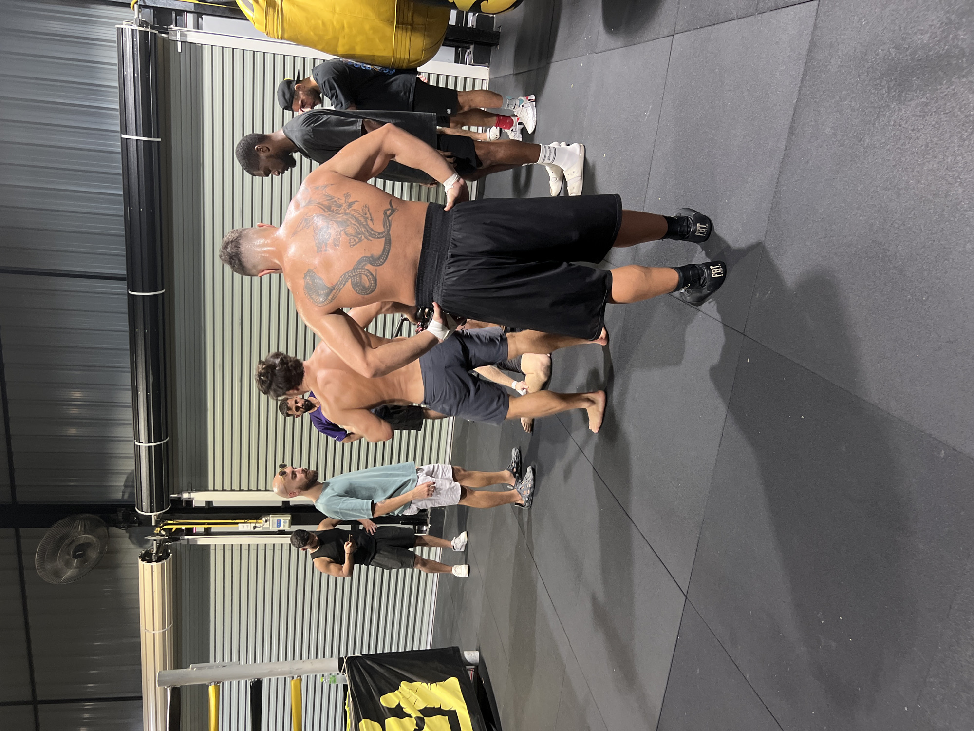 An indoor boxing gym in Chalong, Thailand, is shown with several men present. In the foreground, a man with a large, intricate tattoo of a cobra and dragon on his back is facing away from the camera, wearing black boxing shorts and shoes. To his left, a man in a teal t-shirt and shorts stands with his hands on his hips. Behind them, several other men are gathered. One man in a black tank top and shorts is looking down at his phone. Another man with dark hair, wearing a purple t-shirt, stands with his arms crossed. Further back, two men in athletic wear, one in a black t-shirt and the other in a grey t-shirt, are also present. The gym floor is a dark, rubberized surface, and large, corrugated metal doors are visible in the background. A yellow punching bag hangs to the right of the frame, and a fan is mounted on the ceiling. The lighting suggests it is daytime, with artificial lights illuminating the space.