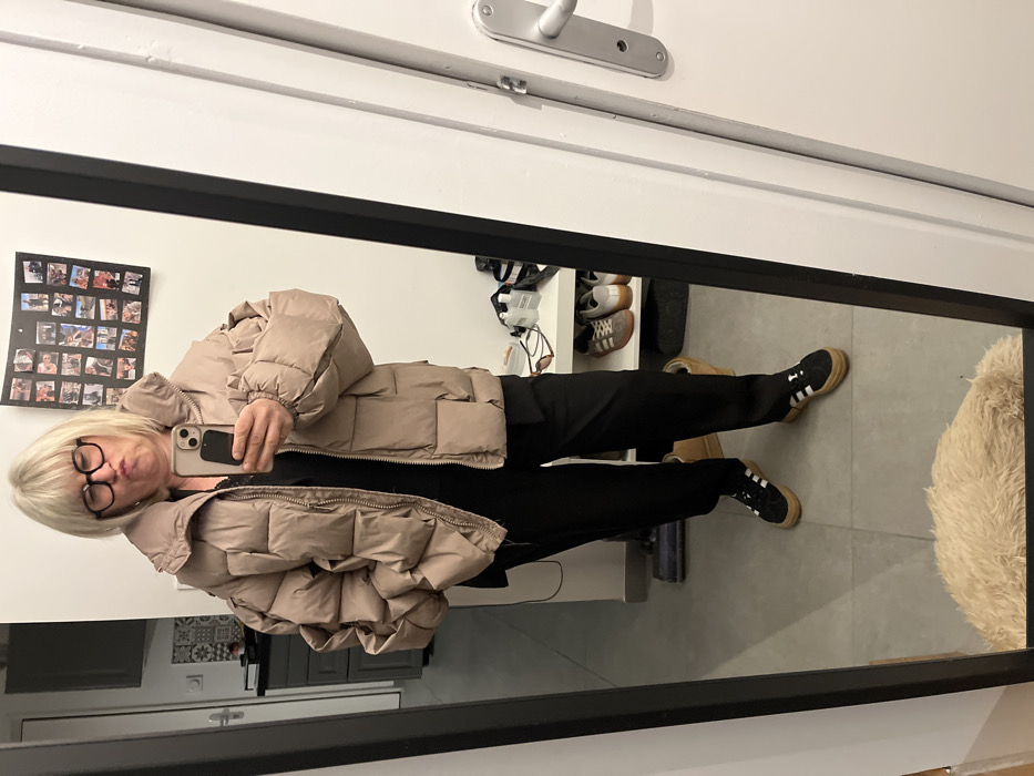 The image depicts a mirror selfie of a person, seemingly a woman, standing indoors. The image is oriented with the person lying horizontally, as if the camera was rotated 90 degrees clockwise during capture or display, but the reflection in the mirror shows them standing upright.
The person has short, light blonde hair with bangs and is wearing black-rimmed glasses. They are making a puckered face towards the camera, which is an iPhone held up by their right hand. The phone case is light pink or beige, and part of the camera lenses are visible.
They are dressed in a thick, light beige or tan puffer jacket with large, distinct horizontal baffles. Beneath the jacket, a black top is visible. They are wearing black trousers that appear to be a relaxed fit, and black sneakers with white stripes on the side and a tan or gum-colored sole, suggestive of an Adidas style like Gazelles or Sambas.
The mirror in which the selfie is taken has a dark, possibly black, frame along the bottom and right side (as oriented in the image). The reflection reveals the room around the person. To their left (our right, if viewing the person upright), a wall is visible with a rectangular collage of multiple small photographs arranged in a grid. Above this, a white door frame and a silver door handle are visible. Further to the person's right (our left), reflected in the mirror, is a shelf or rack with several pairs of shoes, including sneakers and sandals. The floor reflected is a light grey or beige tiled surface, and a fluffy, light-colored (possibly beige or off-white) rug or object is visible in the lower right corner of the mirror's reflection. The lighting suggests an indoor setting, likely with artificial light.