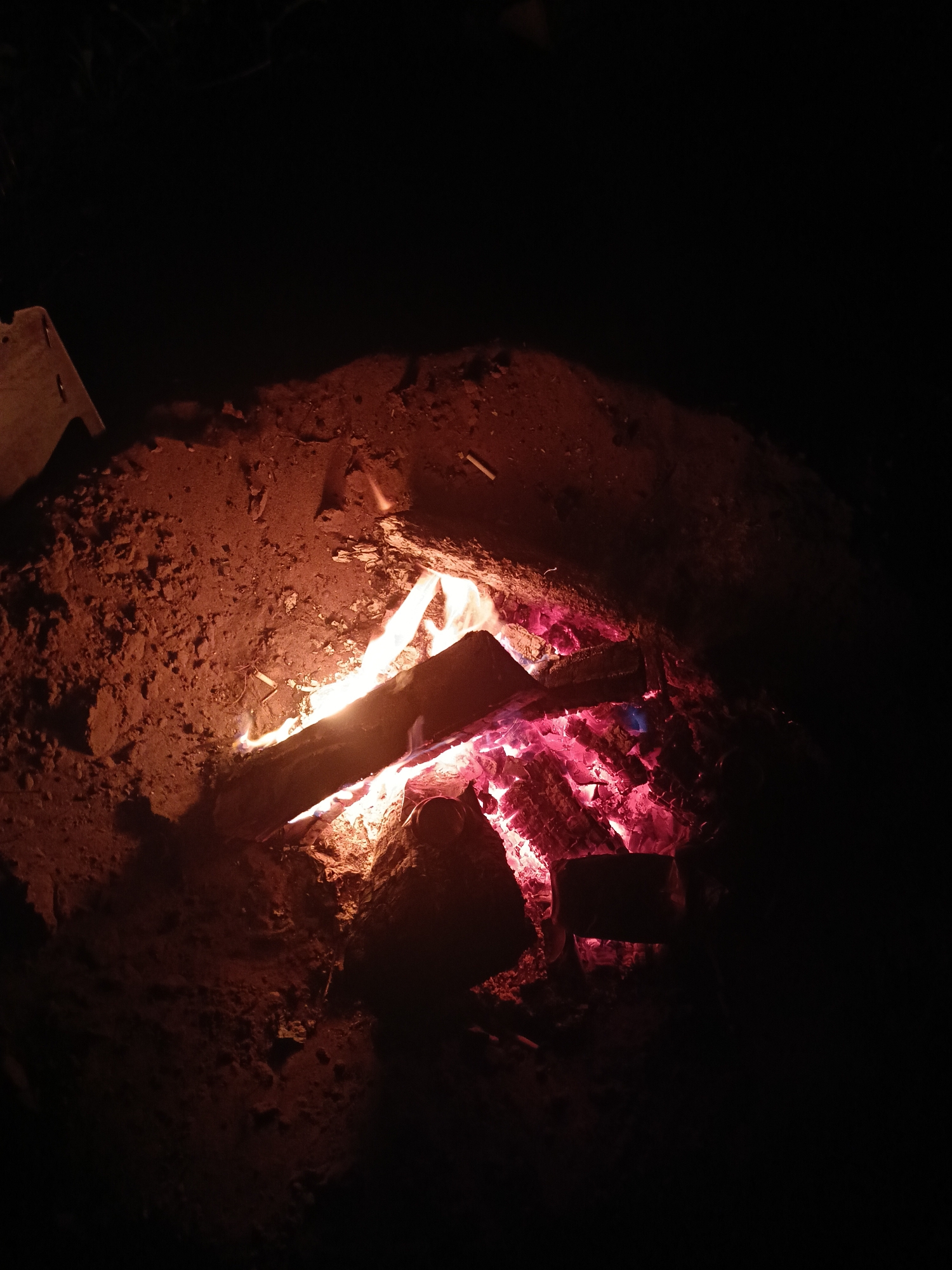 A close-up shot shows a campfire at night. The fire is built within a shallow pit surrounded by dark, reddish-brown dirt. Burning logs and embers glow with orange, yellow, and red light. The darkness around the fire obscures details, suggesting a remote location. There are no visible people. The city name, Noviy, Russia, is not evident in the image.