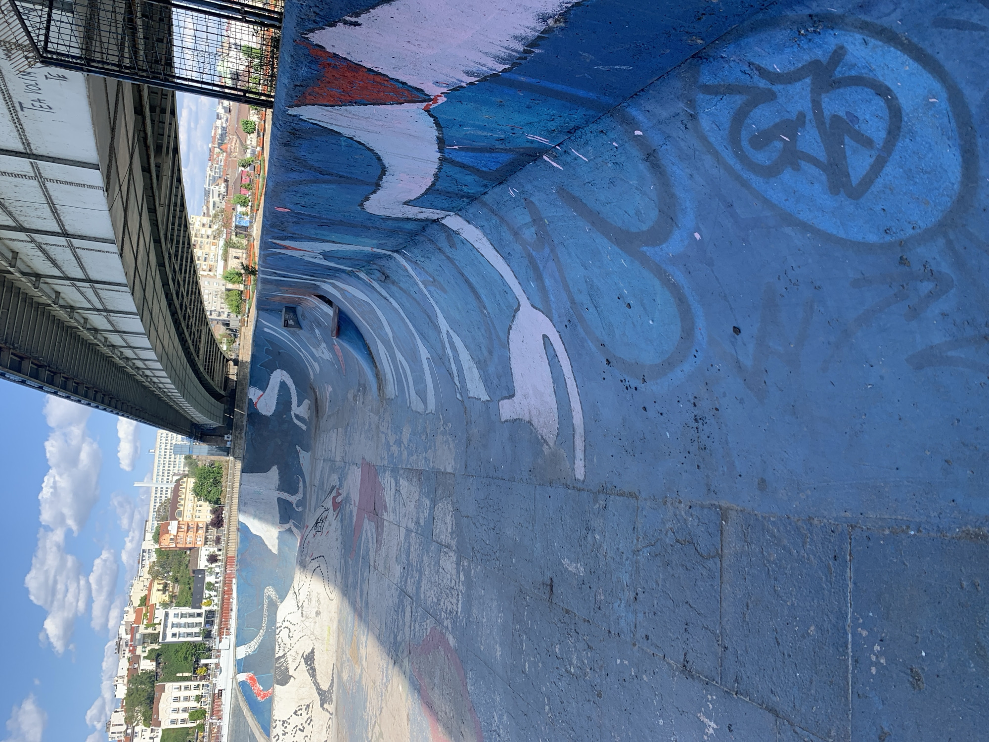 The image shows a segment of a graffiti-covered wall or ramp, possibly within an urban park setting in Belgrade, Serbia. In the background, there is a view of a city skyline with buildings, trees, and a blue sky with clouds. An elevated structure, possibly a bridge or walkway, is visible above the wall, partially obscuring the sky. The wall itself is primarily blue, adorned with various white and grey graffiti designs. The graffiti appears to be abstract shapes and symbols. The lighting suggests it is daytime, and the photo is taken from a slightly elevated position.