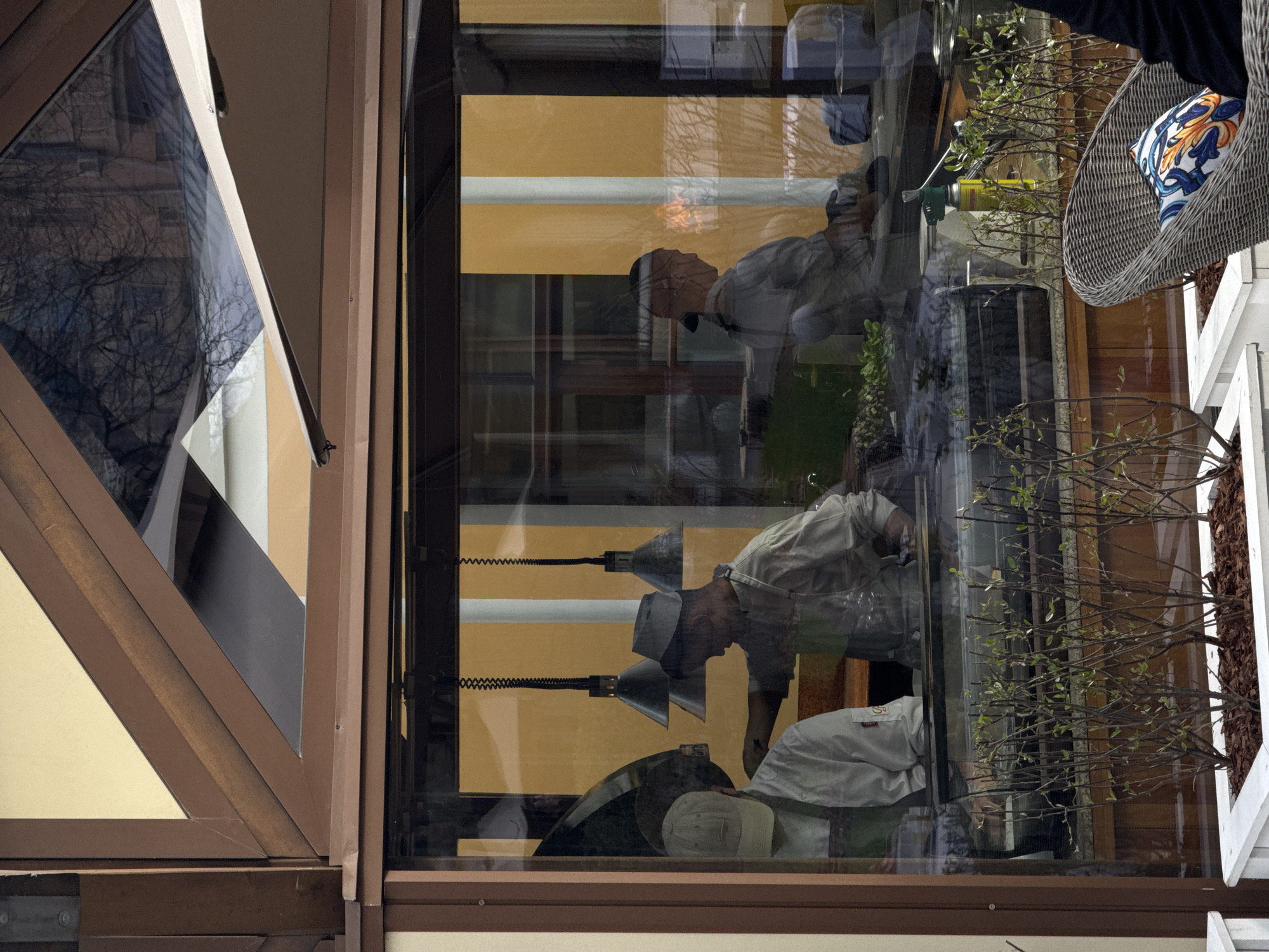 The image shows a scene reflected in a glass window, potentially the exterior of a building. The location is suggested to be Moscow, Russia, based on the architectural style of the building's window frame.
The reflection includes:
* People: At least two individuals are reflected, possibly workers.
* Objects: Work lamps, plants, and what appears to be a wicker chair with a patterned cushion are present.
* Environment: The reflection shows elements of an interior setting with visible walls and other furnishings. The window frame is made of brown wood.
Due to the reflection, the details are somewhat obscured, but the image captures a snapshot of activity within a specific architectural setting.